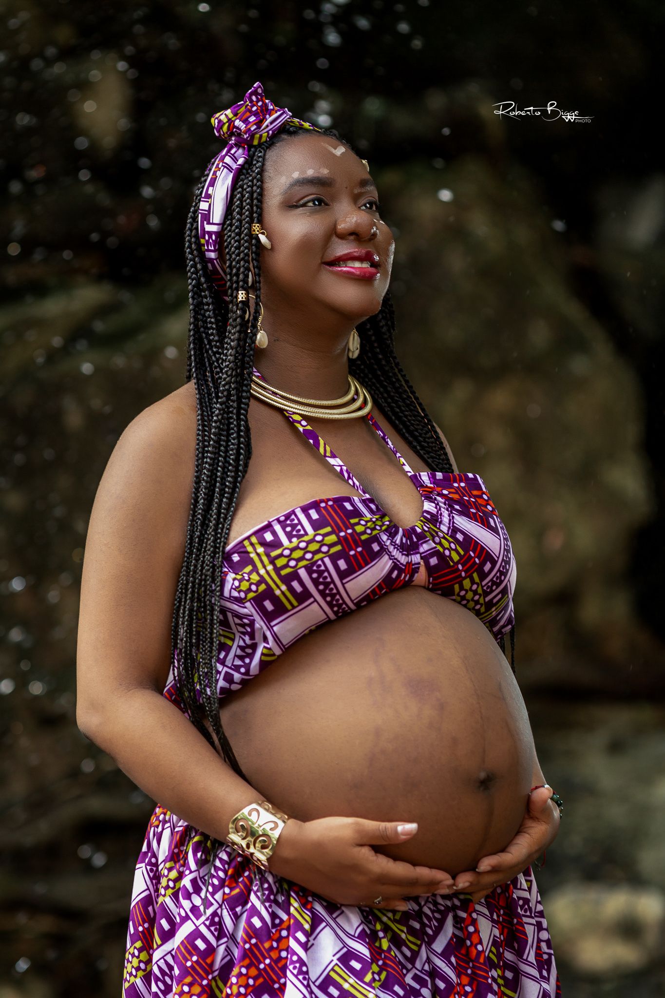 Expecting mother in vibrant purple and pink patterned dress with braided hair poses outdoors during maternity photoshoot.