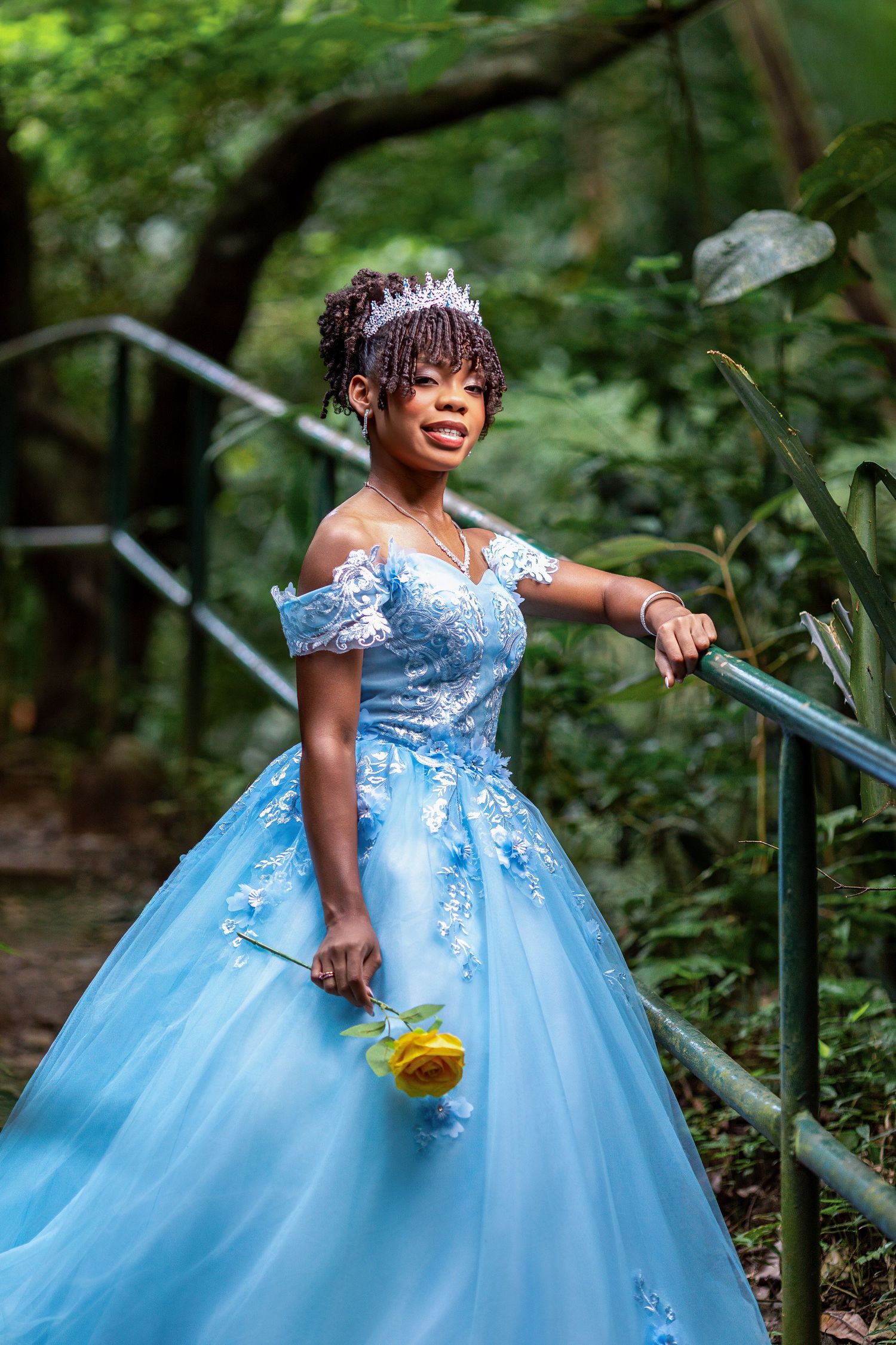 Young Black woman in blue ball gown with floral lace, tiara, holding yellow rose on garden staircase.