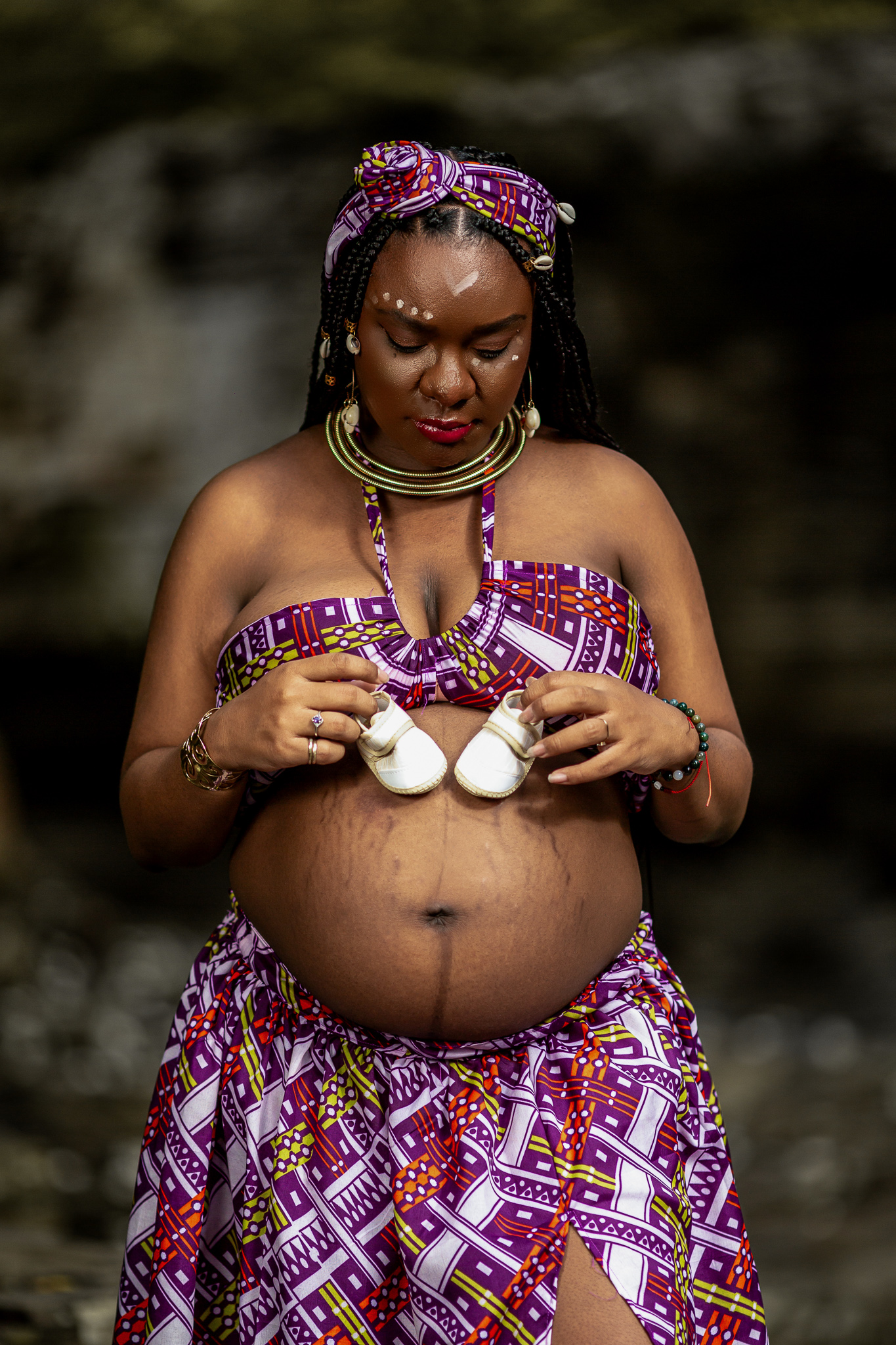 Maternity photo in African print dress with baby shoes against natural backdrop.