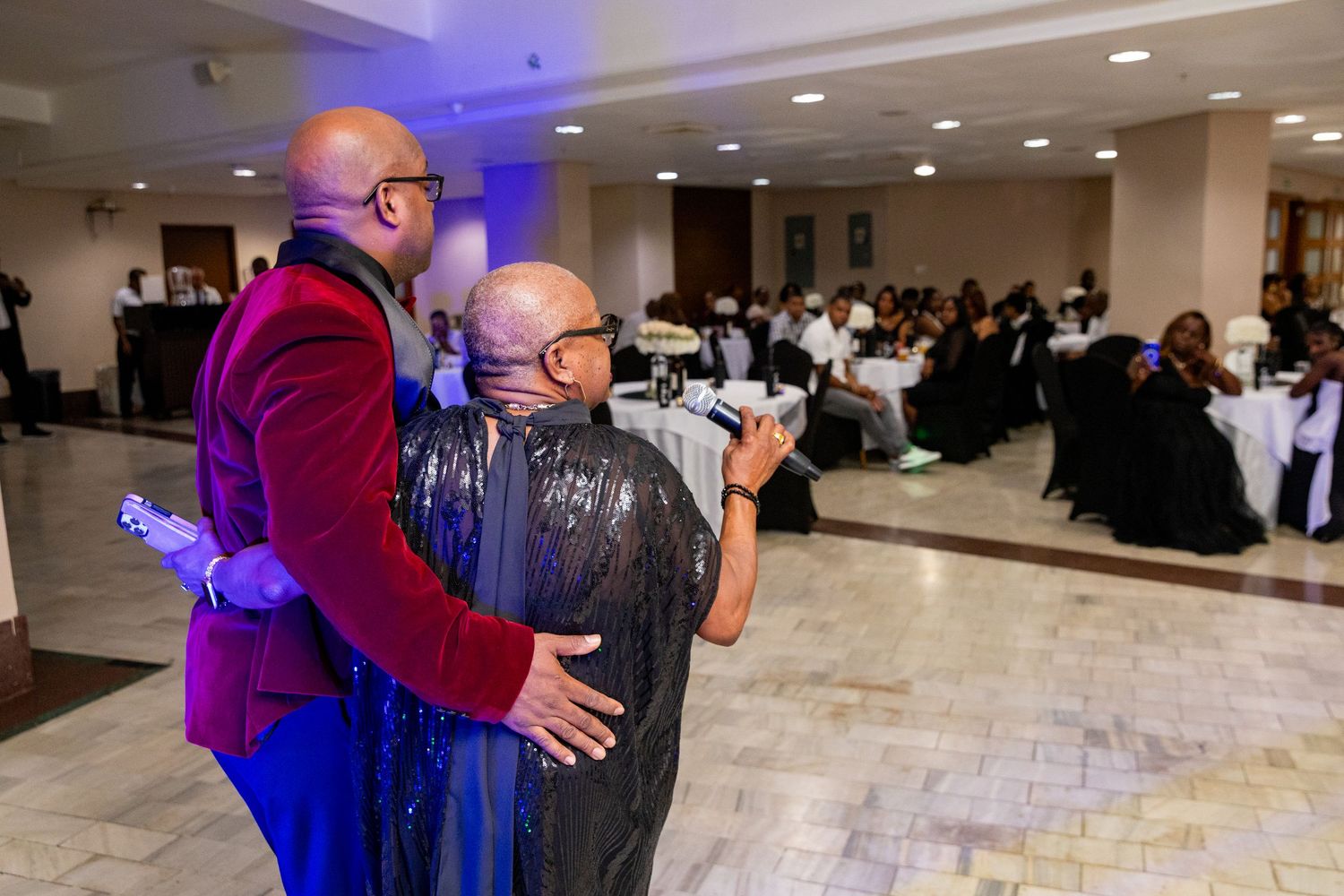 Two elegantly dressed guests share a warm embrace on the dance floor at a formal banquet event.
