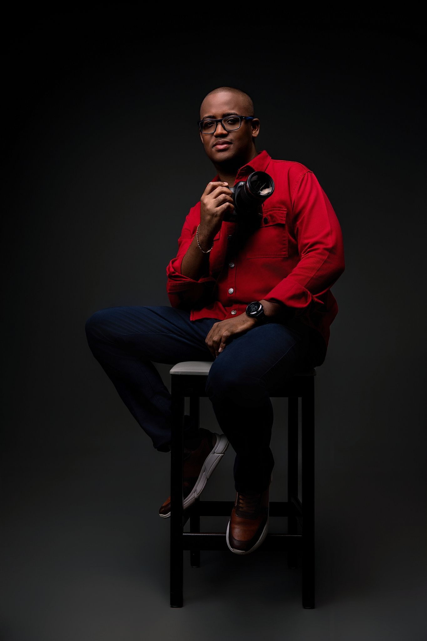 A photographer in a red button-down shirt and glasses sits on a black stool holding a camera against a dark background.