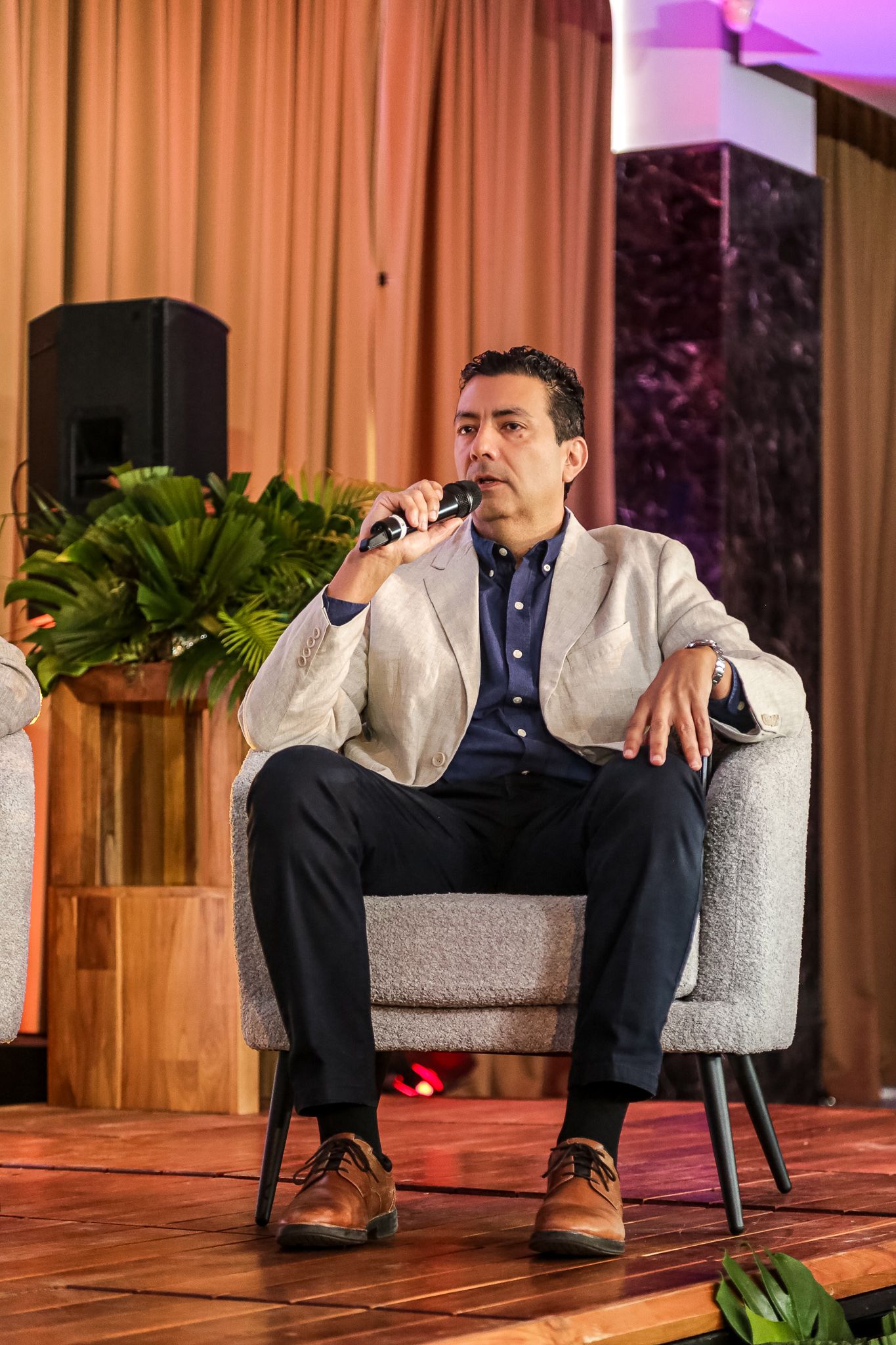 A person in business attire sits thoughtfully in a gray armchair on a wooden stage with pink lighting.