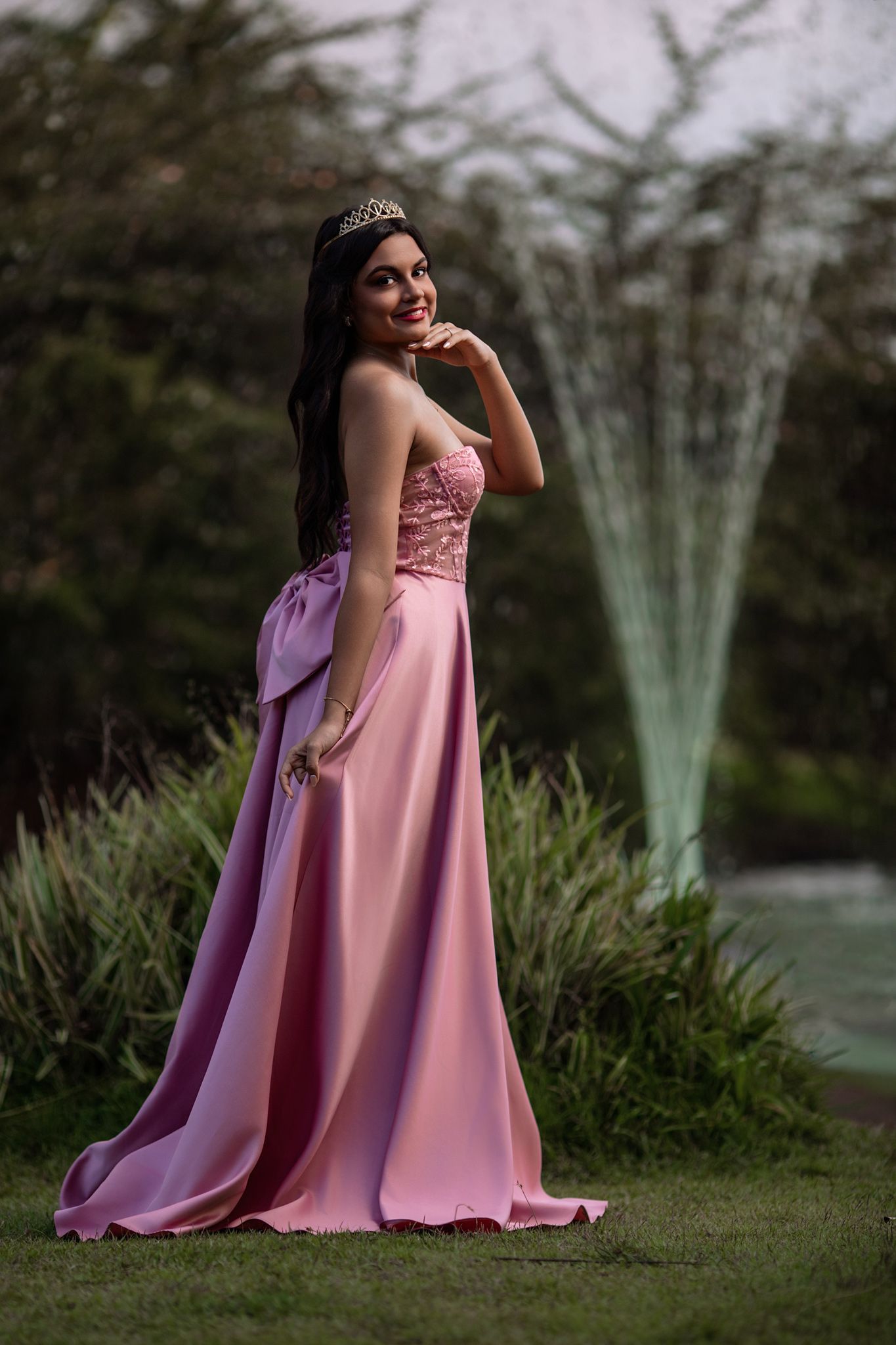 A person in a flowing pink formal gown poses gracefully outdoors near a fountain and greenery.