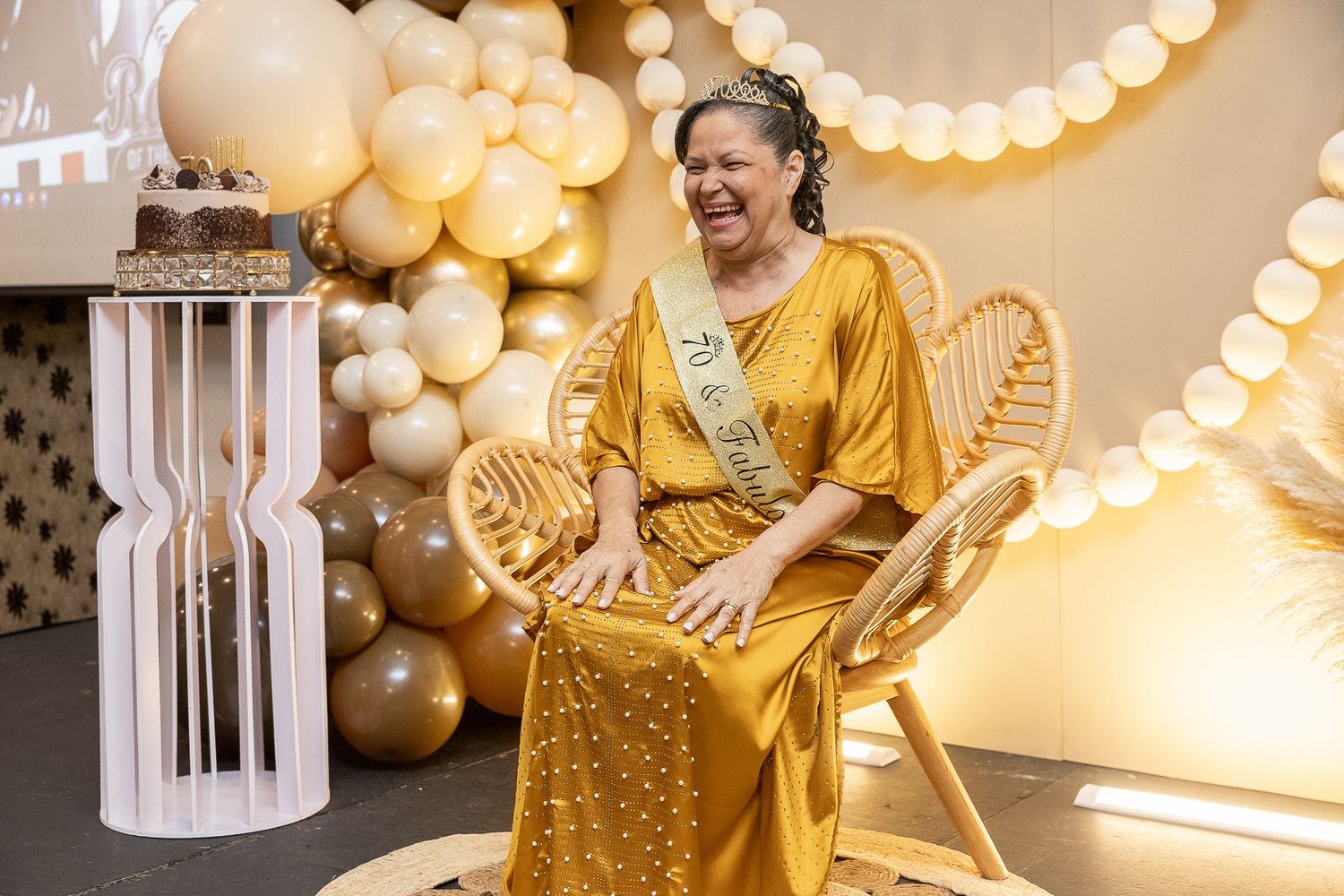 Woman in gold dress laughing joyfully at a golden birthday party with balloon decorations and a sash.