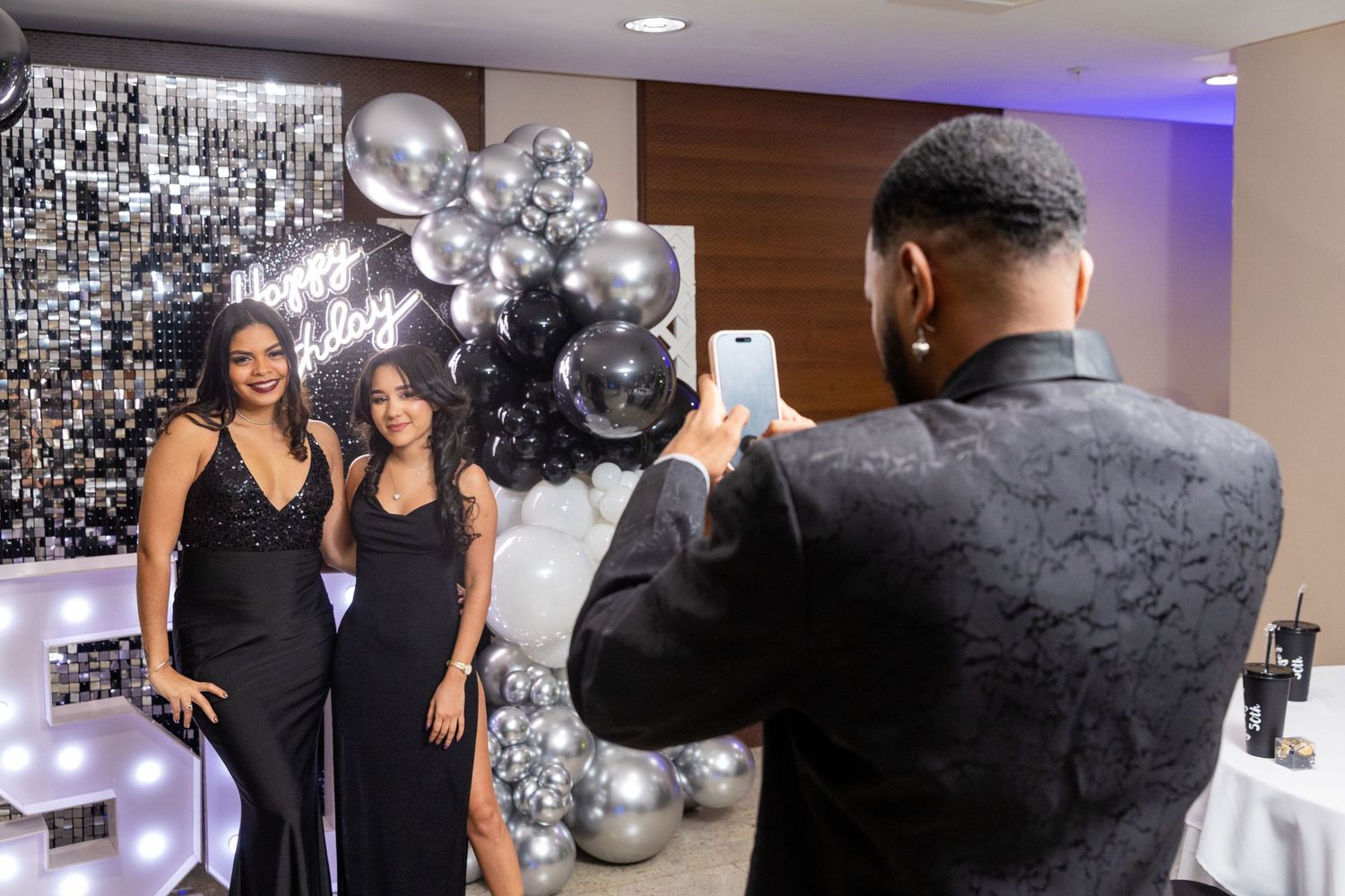 A man photographs two women in black dresses posing at a glamorous birthday party celebration.