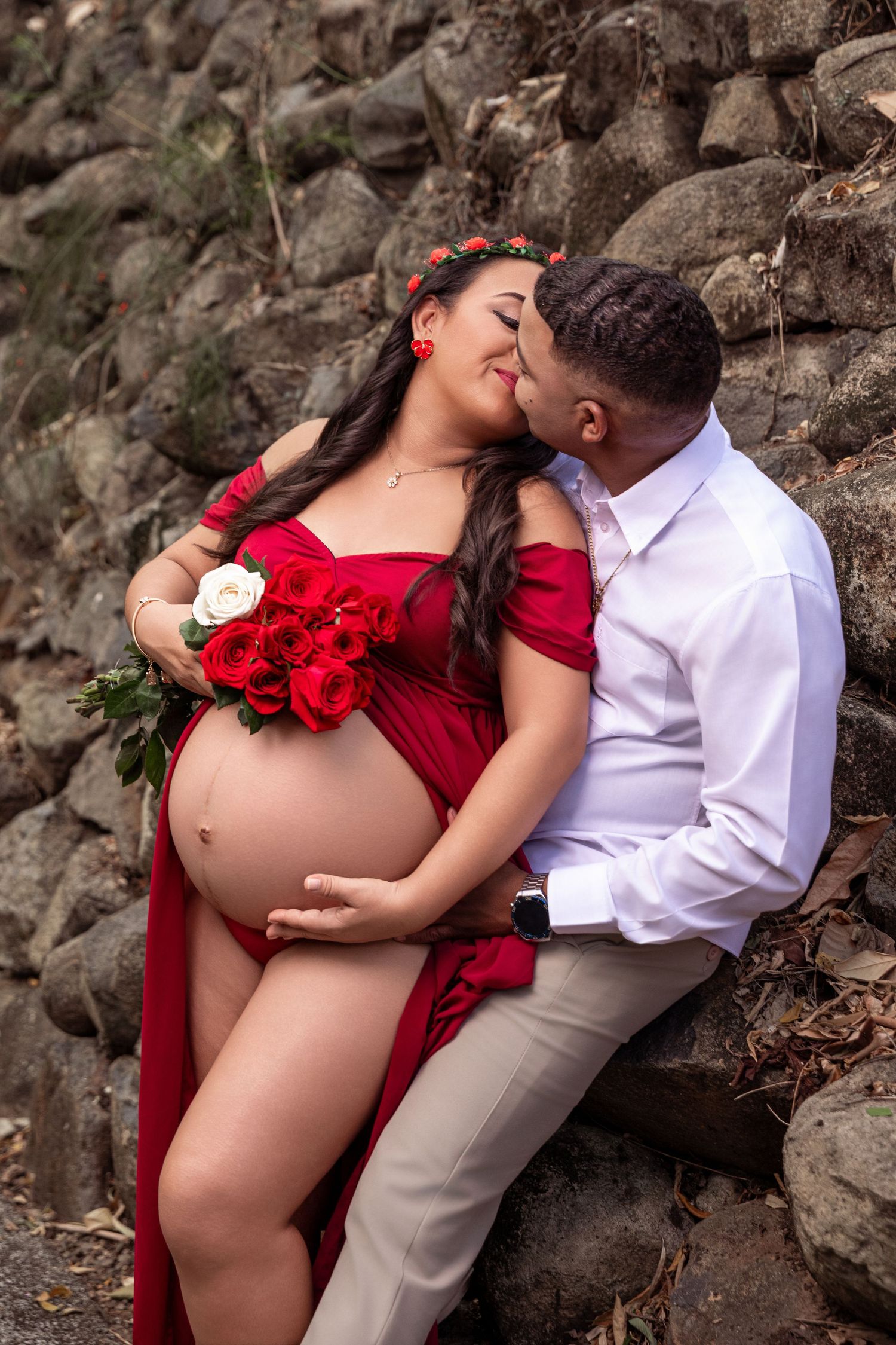 Pregnant woman in red dress holding roses, kissed by partner against stone wall in romantic maternity photo shoot.