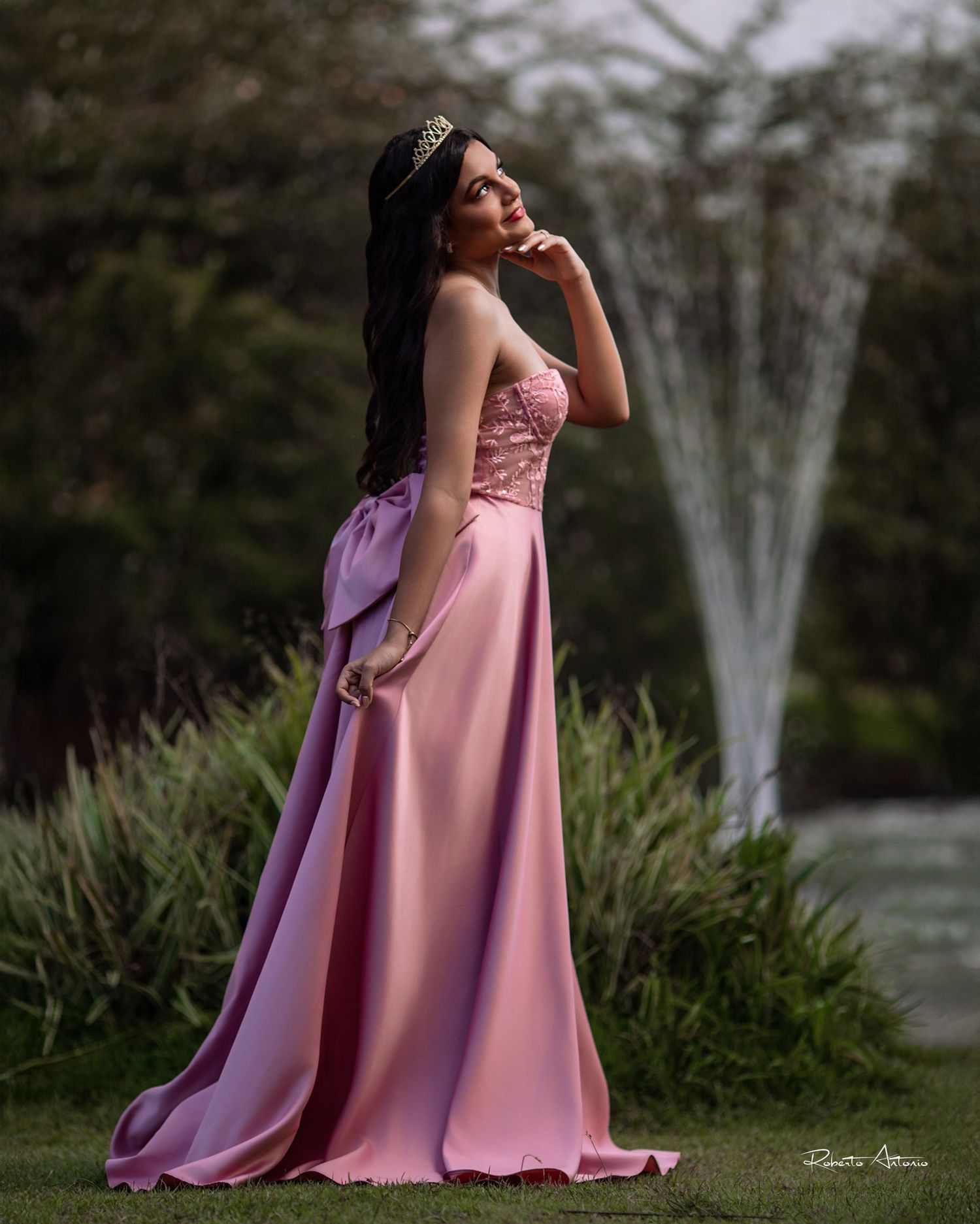 A person in a flowing pink evening gown poses gracefully in a garden setting with dramatic lighting at dusk.