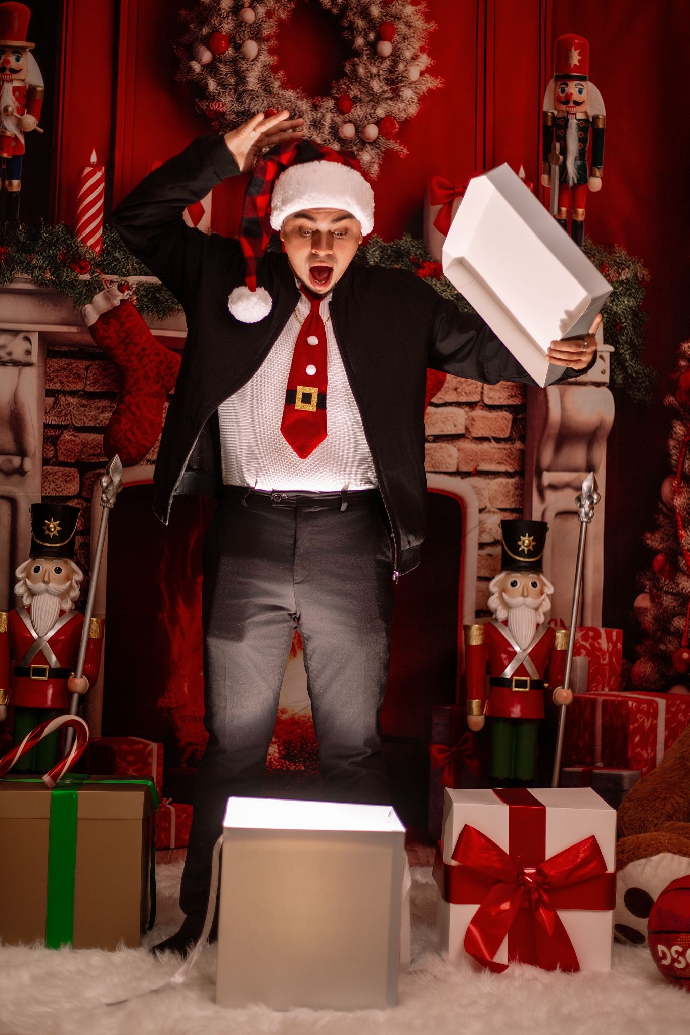 Person in Santa hat and suit vest opens gifts against festive red holiday backdrop with Christmas decorations.