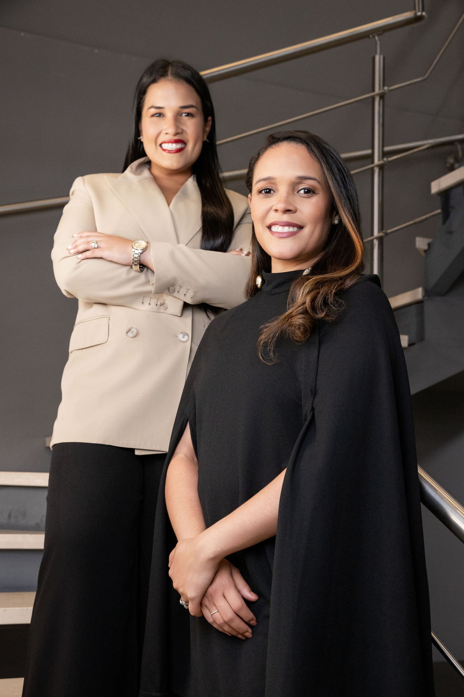 Two professional women in elegant attire smile confidently on modern stairs, one in beige blazer, one in black cape dress.