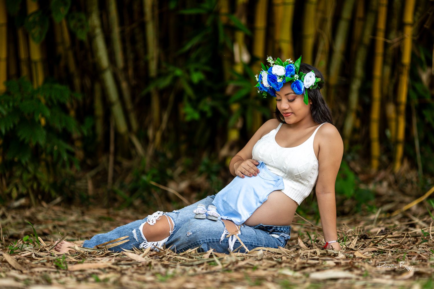 Pregnant woman in ripped jeans and white tank top relaxes in bamboo forest with blue flower crown during maternity shoot.