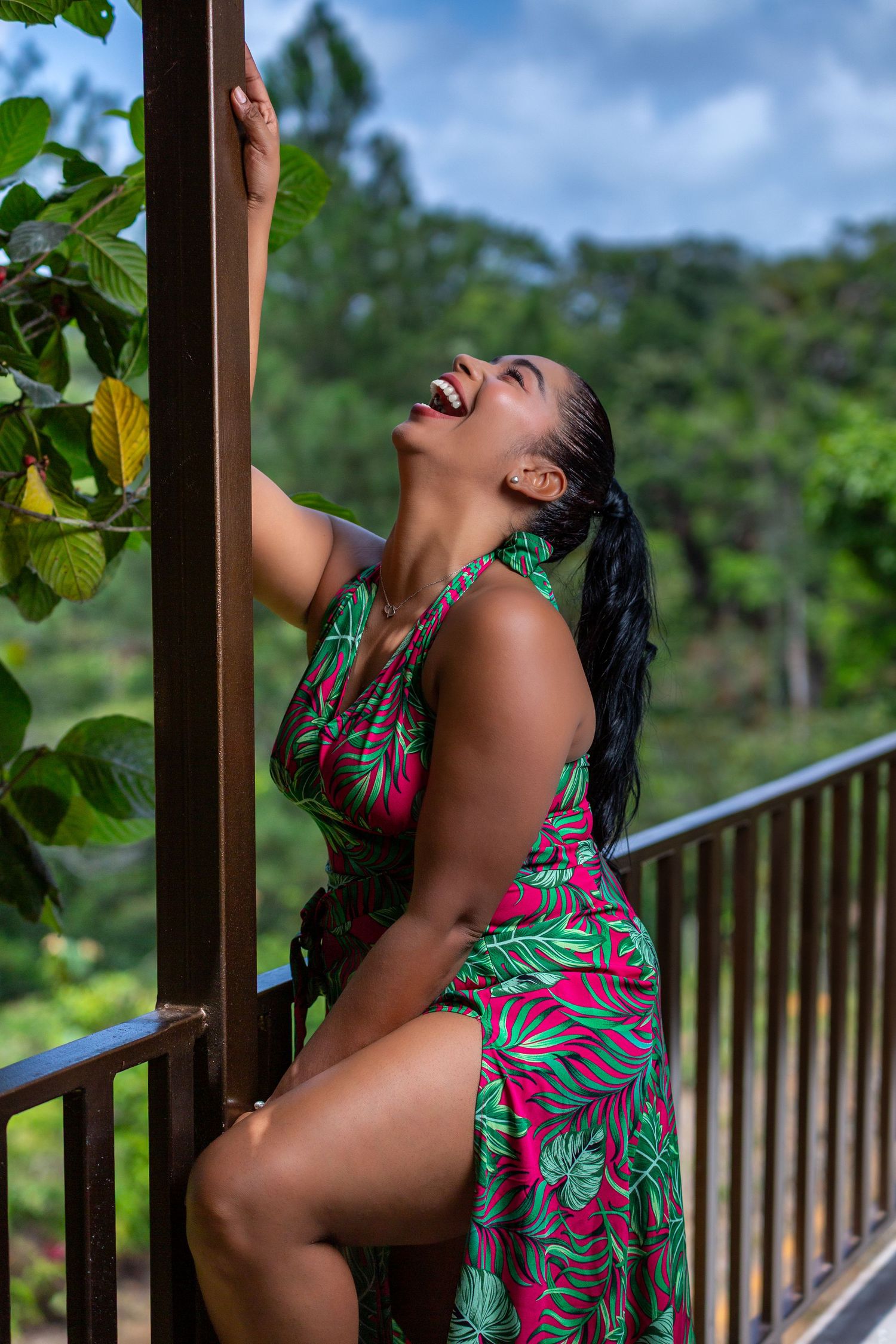 Model in tropical print dress poses expressively on outdoor deck surrounded by greenery.