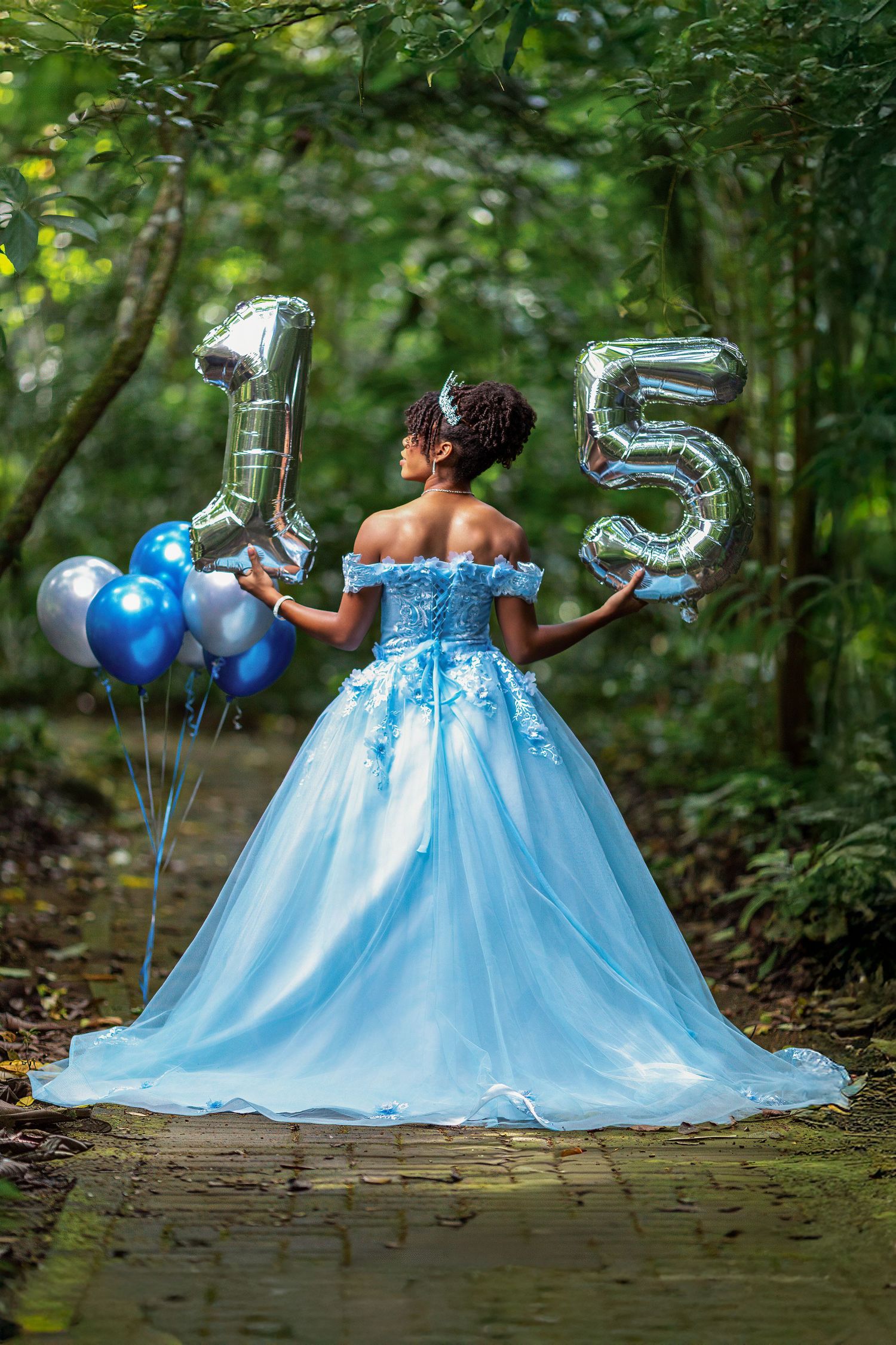 Young woman in blue quinceañera gown holds silver number 15 balloons in lush green garden path.