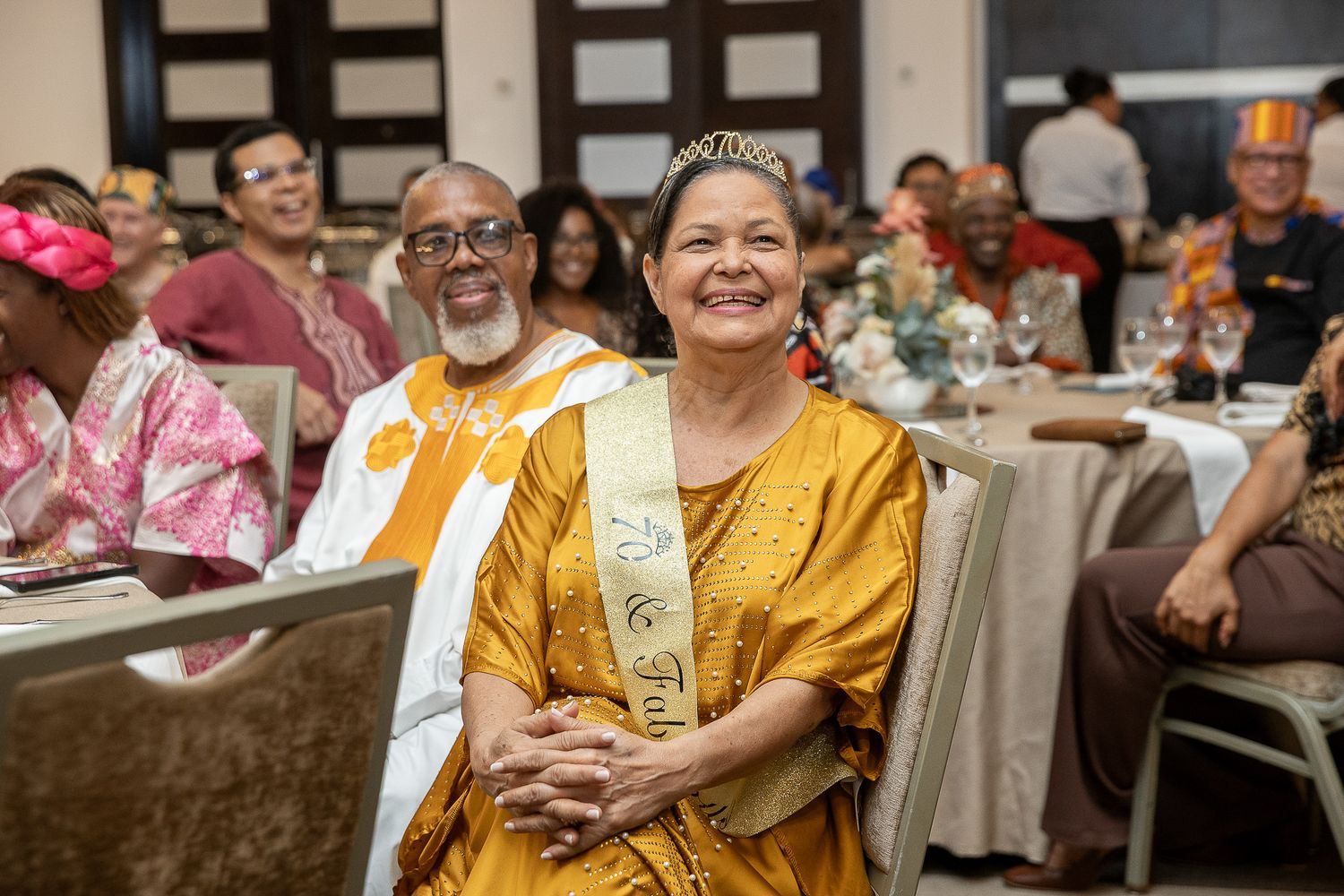 Woman in golden dress and tiara sash smiles at formal banquet dinner event with guests in traditional African attire.