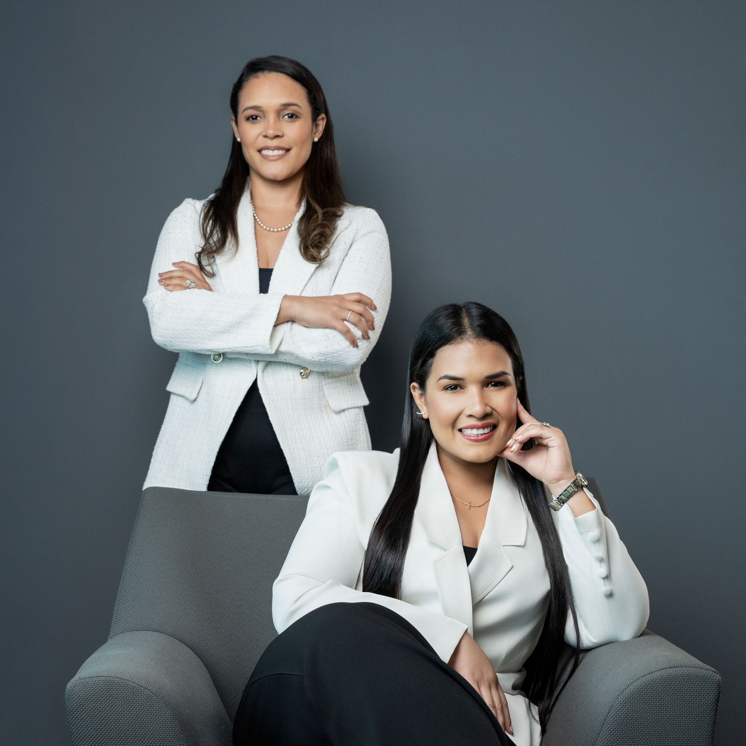 Two professional women in white blazers pose confidently in a modern studio setting.