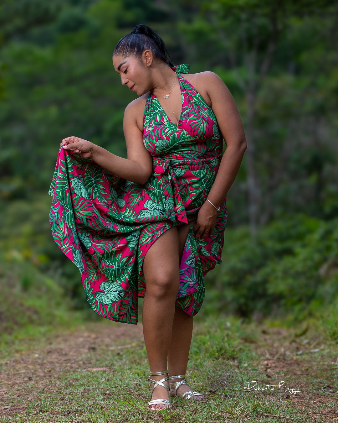 A person wearing a vibrant tropical print halter dress poses outdoors on a dirt path surrounded by greenery.