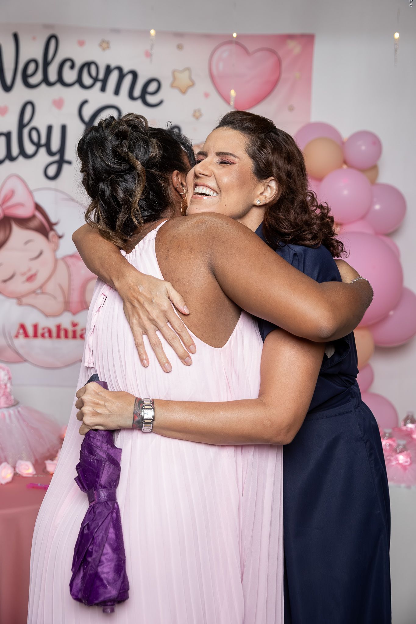 Two women embracing warmly at a baby shower decorated with pink balloons and a Welcome Baby girl banner.
