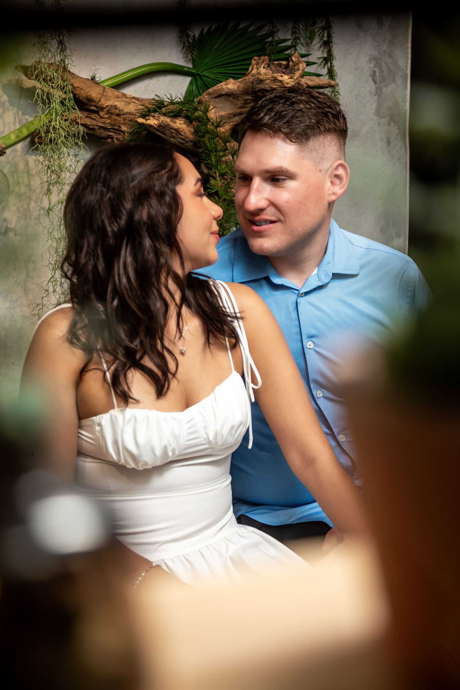 A couple shares an intimate moment together outdoors, with her in white and him in blue against a natural backdrop.