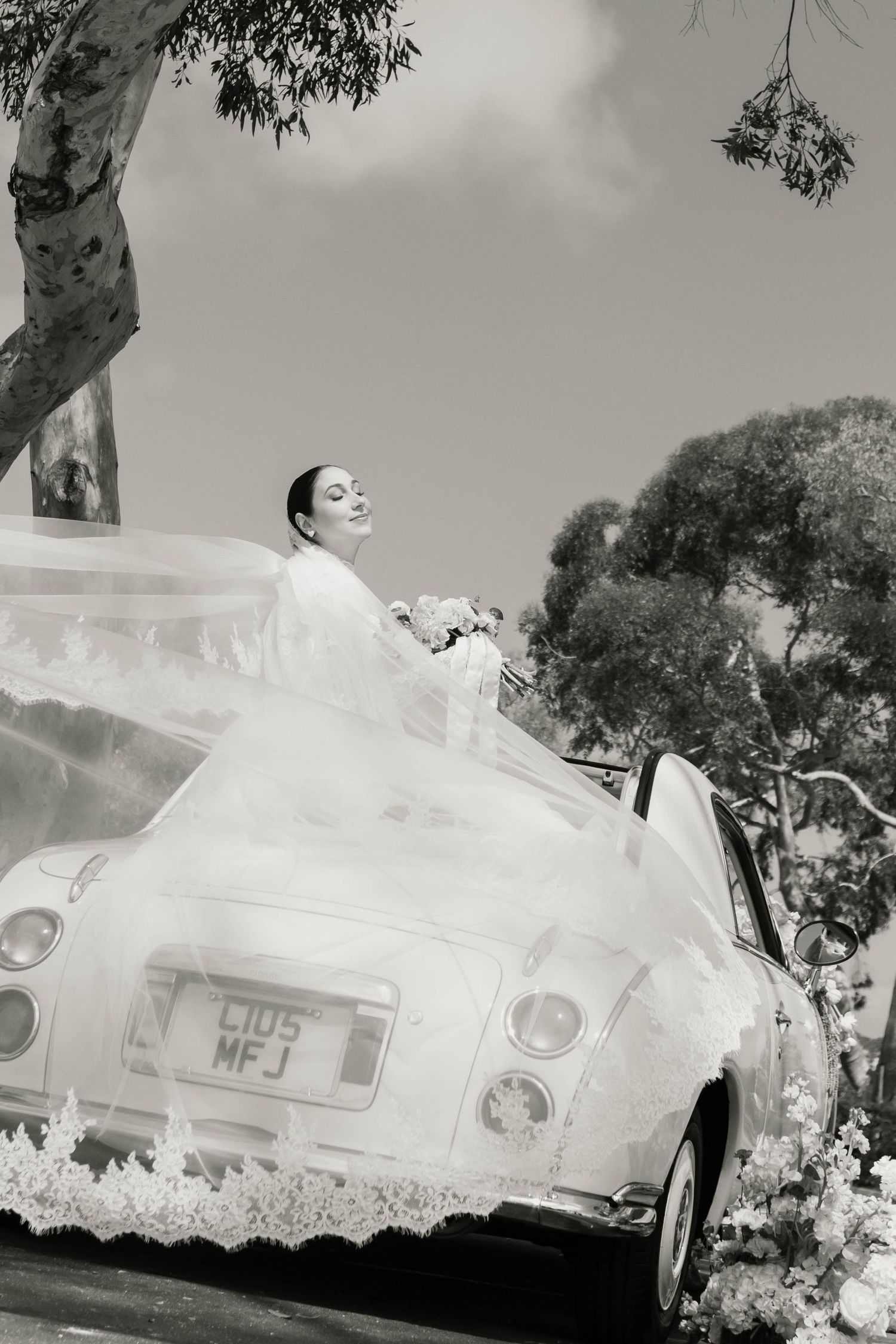 Bride in flowing white gown standing in vintage convertible car decorated with flowers, black and white wedding photo.