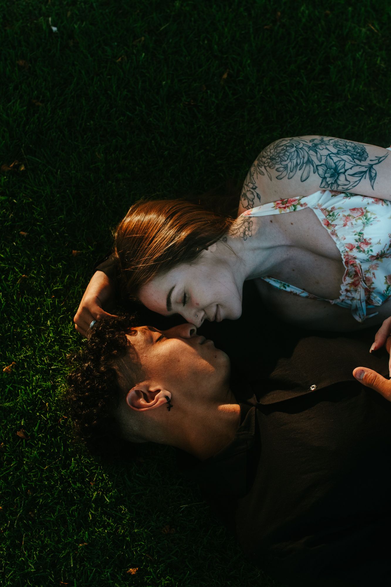 Couple lying on lush green grass sharing an intimate moment, woman with floral tattoo smiling closely.