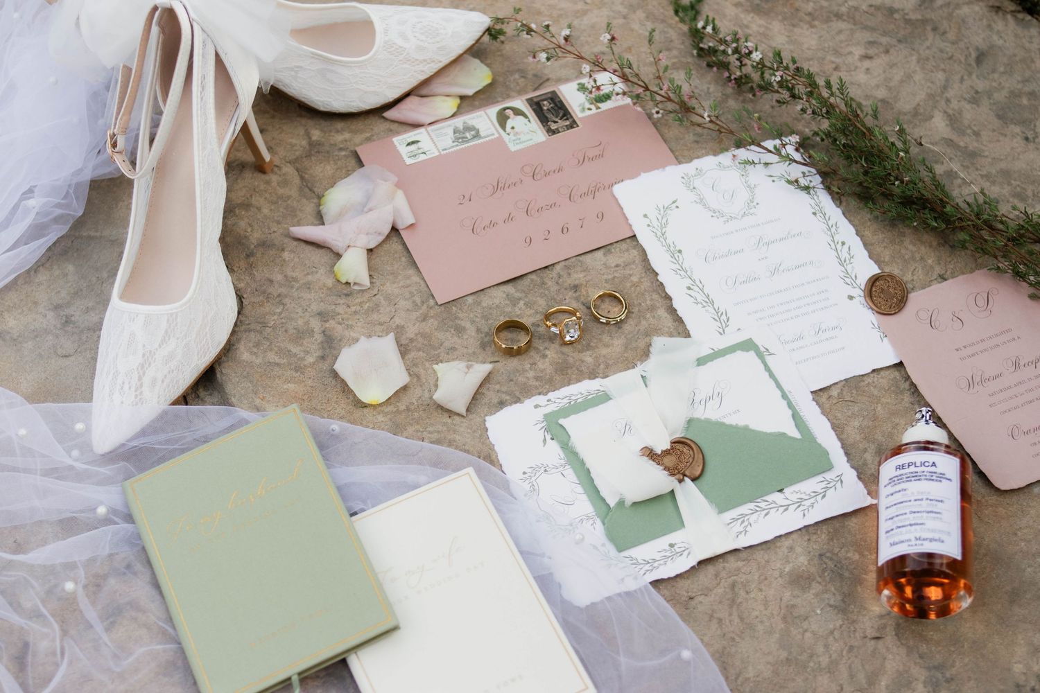 Flat lay of wedding stationery, rings, bridal shoes, and accessories in sage green and blush.