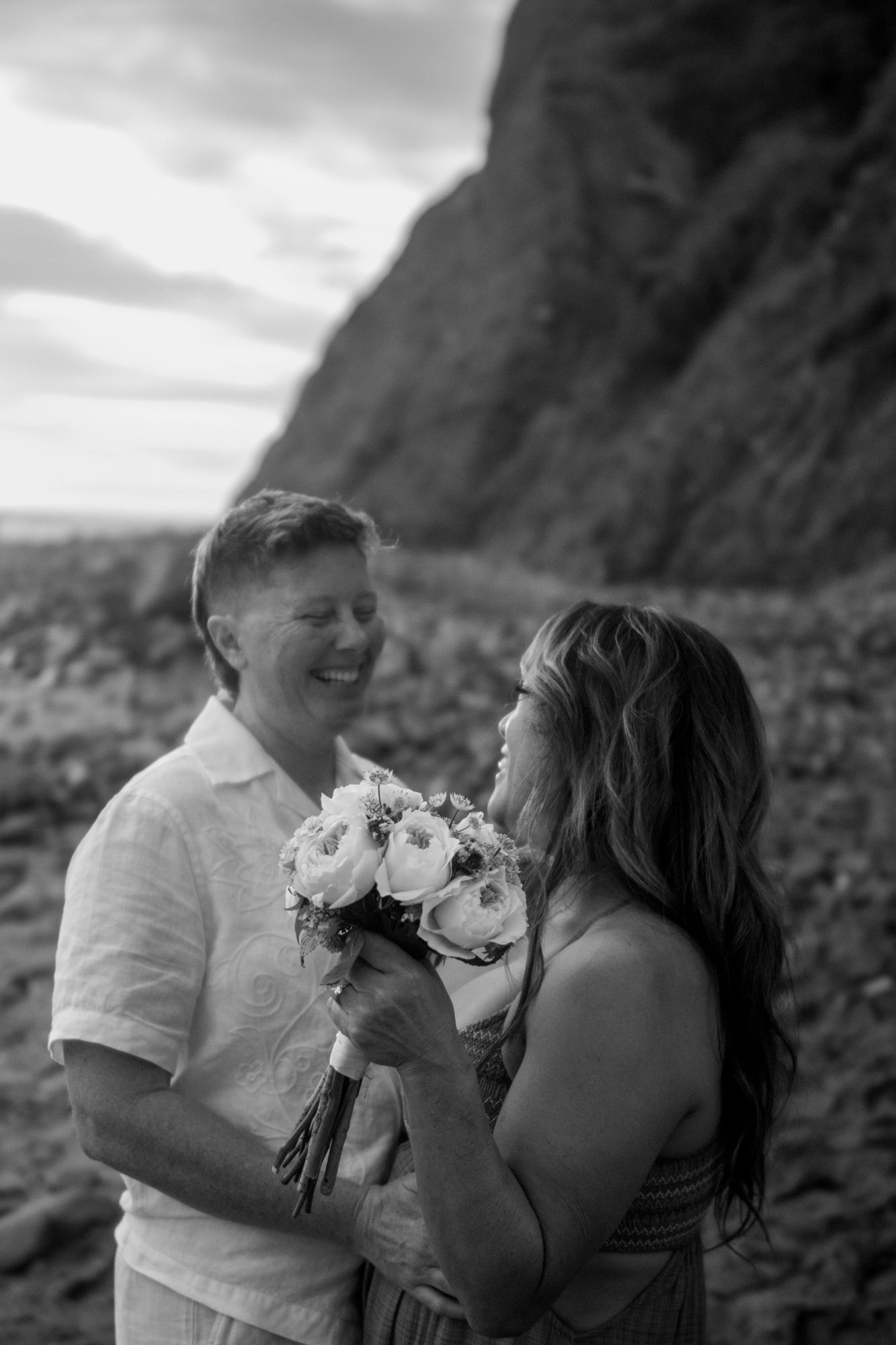 Couple smiling at each other holding bridal bouquet on rocky beach in black and white.