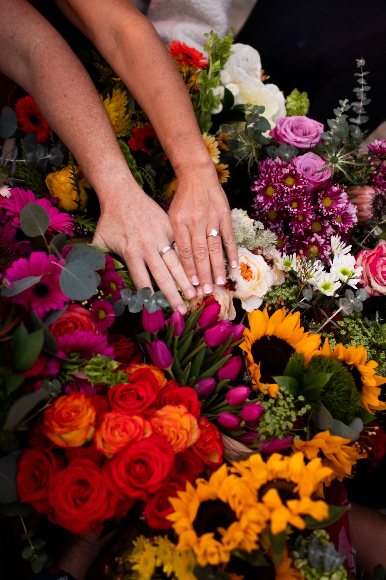 Newlyweds showing wedding rings over colorful floral bouquets of roses and sunflowers.