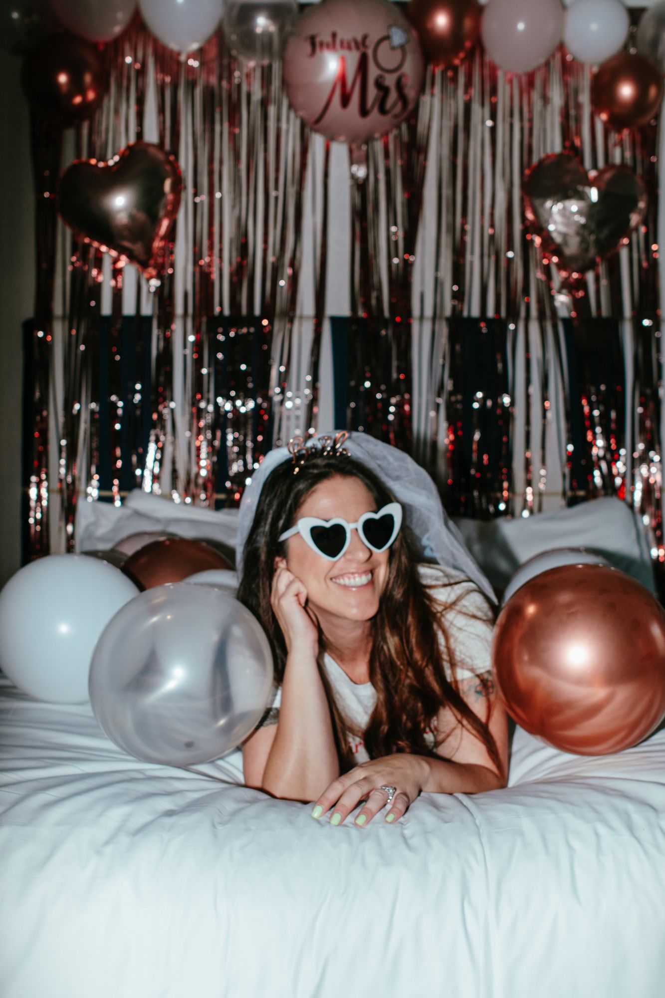 Smiling bride-to-be wearing heart sunglasses and veil lying on bed surrounded by rose gold and silver balloons at bachelorette party.