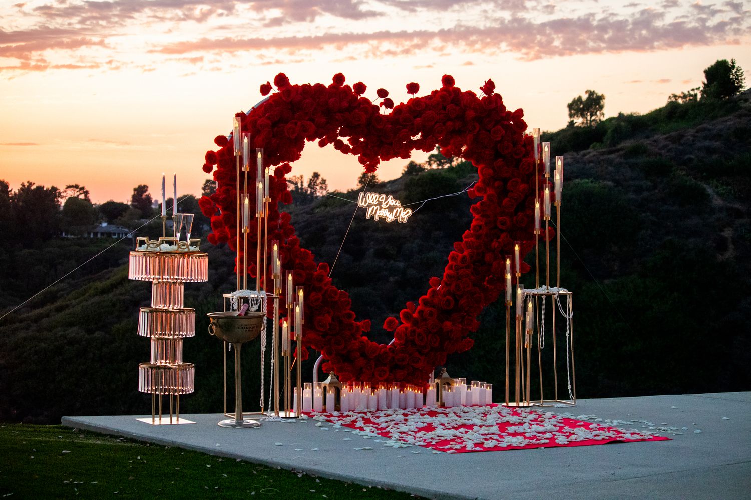 Large red rose heart-shaped floral arch with candles and rose petals at sunset outdoor wedding ceremony.