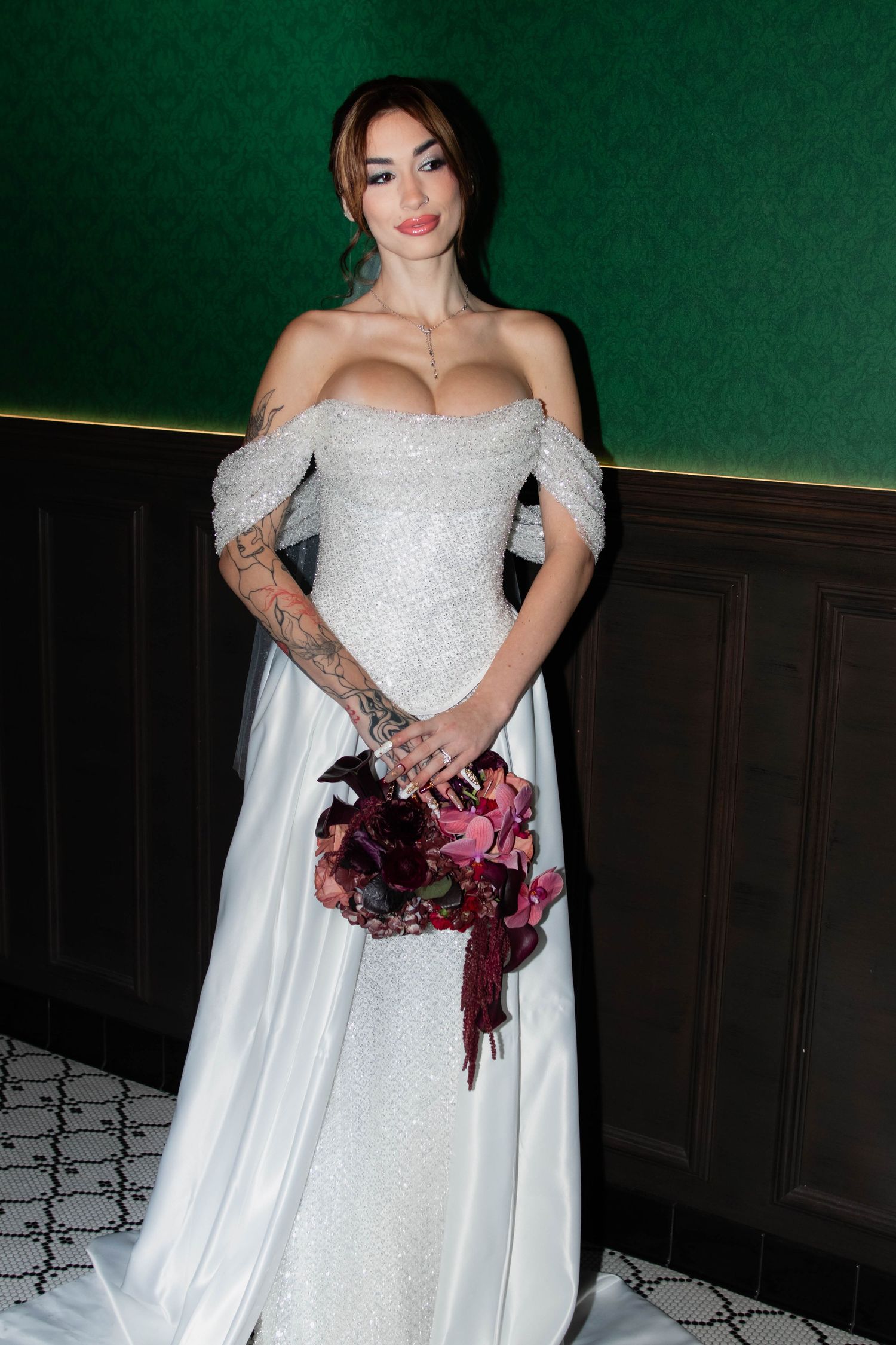Tattooed bride in off-shoulder white gown holds burgundy and pink bouquet against green wall backdrop.