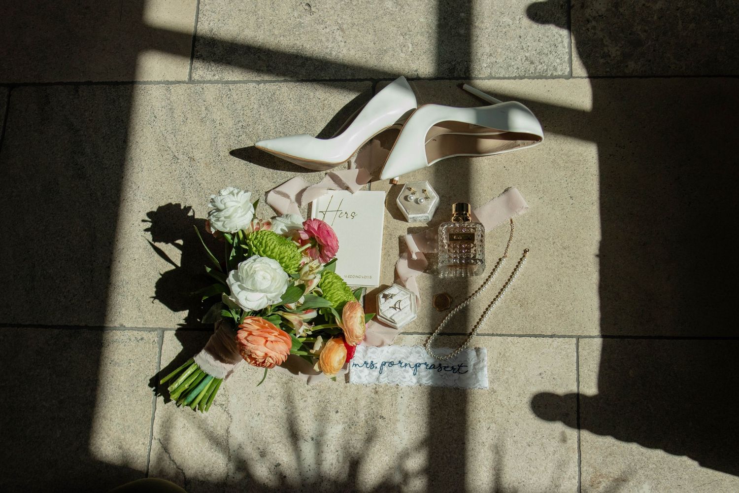 Bridal flat lay with white heels, floral bouquet, perfume bottle, and wedding accessories on stone floor.