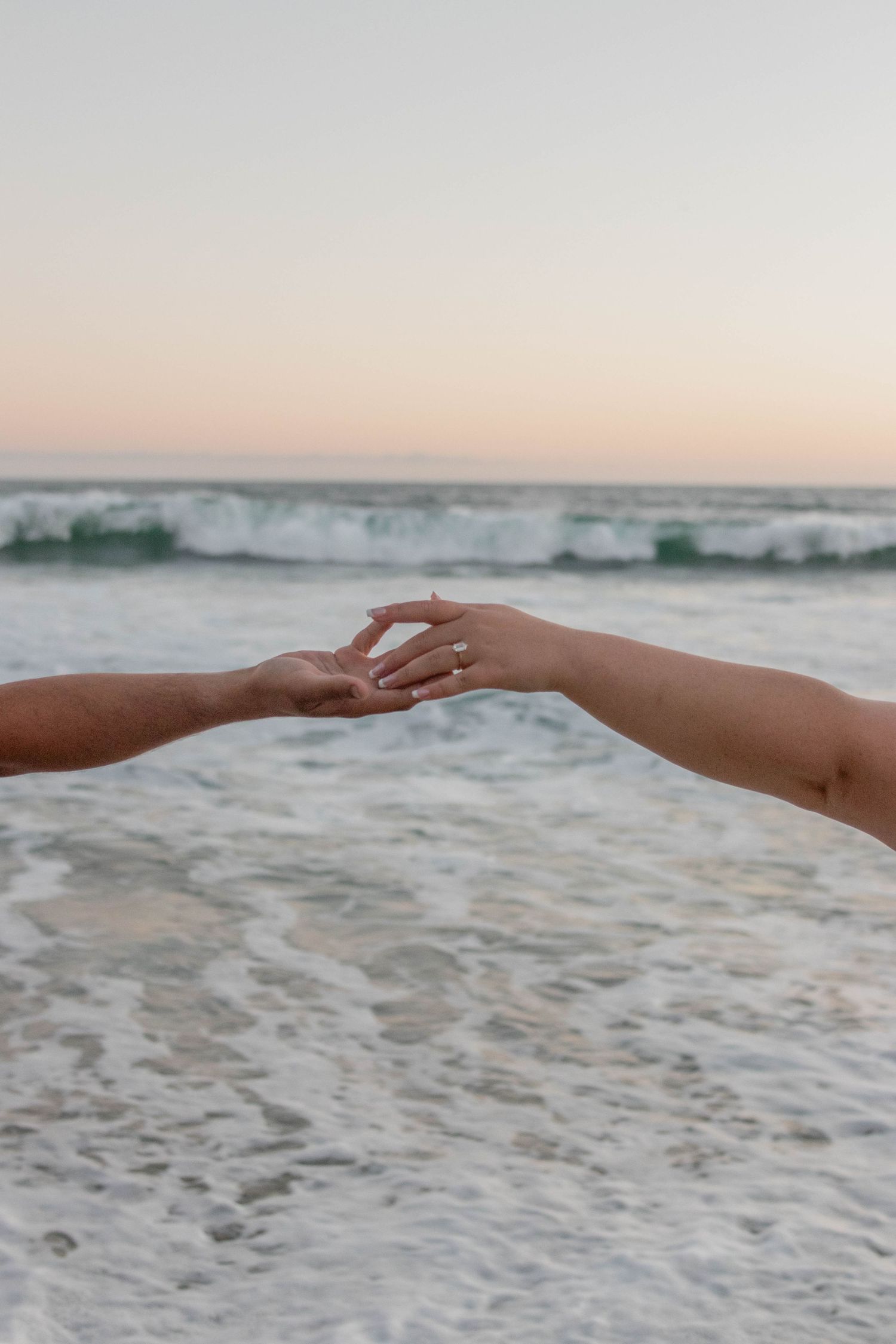 Two hands gently touching fingertips at a serene beach during sunset with soft ocean waves in the background.