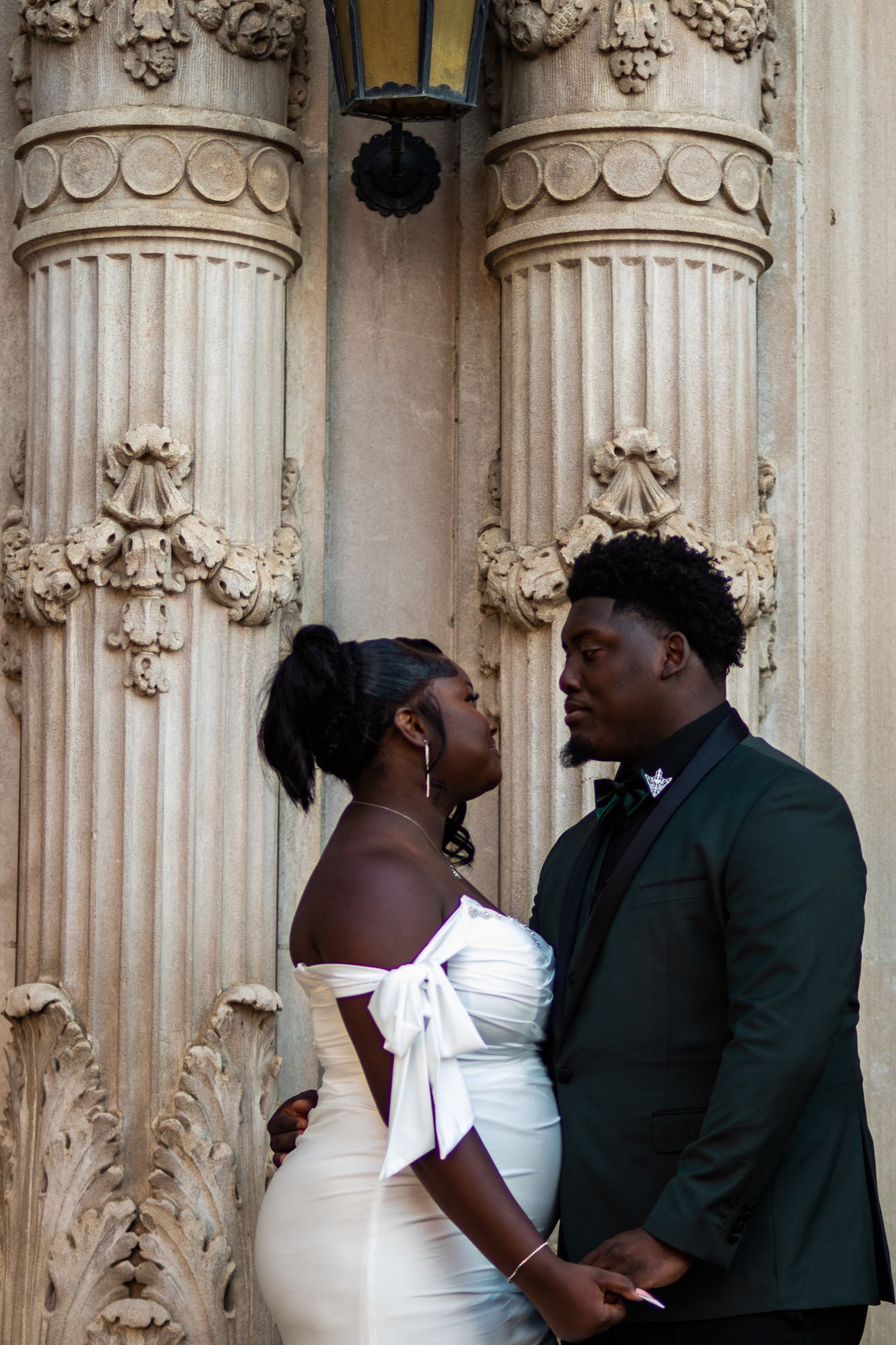 Black couple in wedding attire gazing at each other before ornate stone columns.