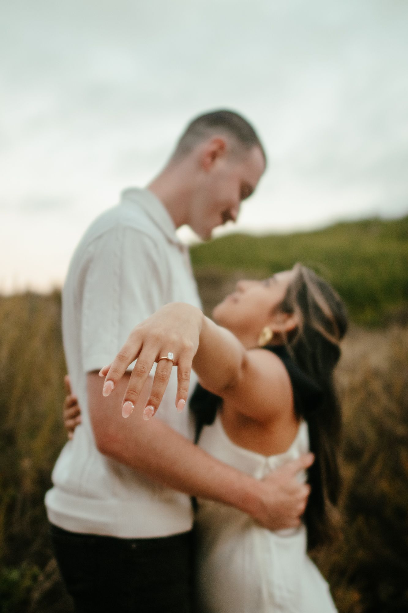Engaged couple embracing in a field displaying diamond engagement ring toward camera at dusk.