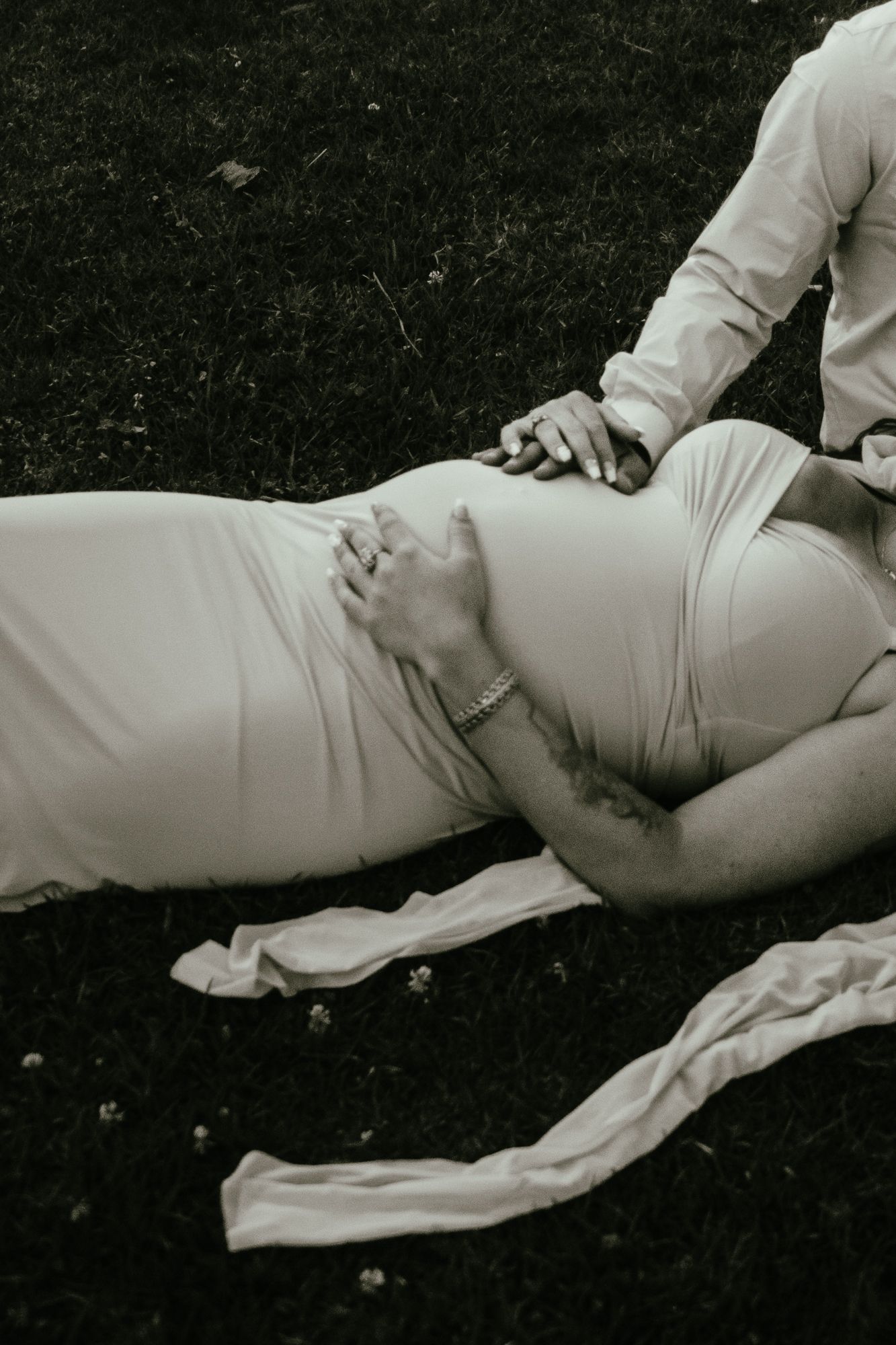 Black and white maternity photo of pregnant woman in white dress lying on grass with partner.