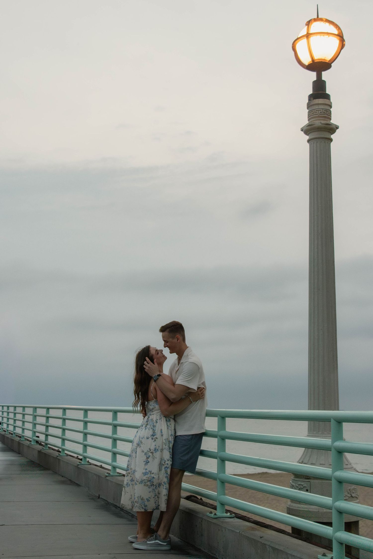 Couple embracing romantically on a pier at dusk under a glowing street lamp with ocean views.