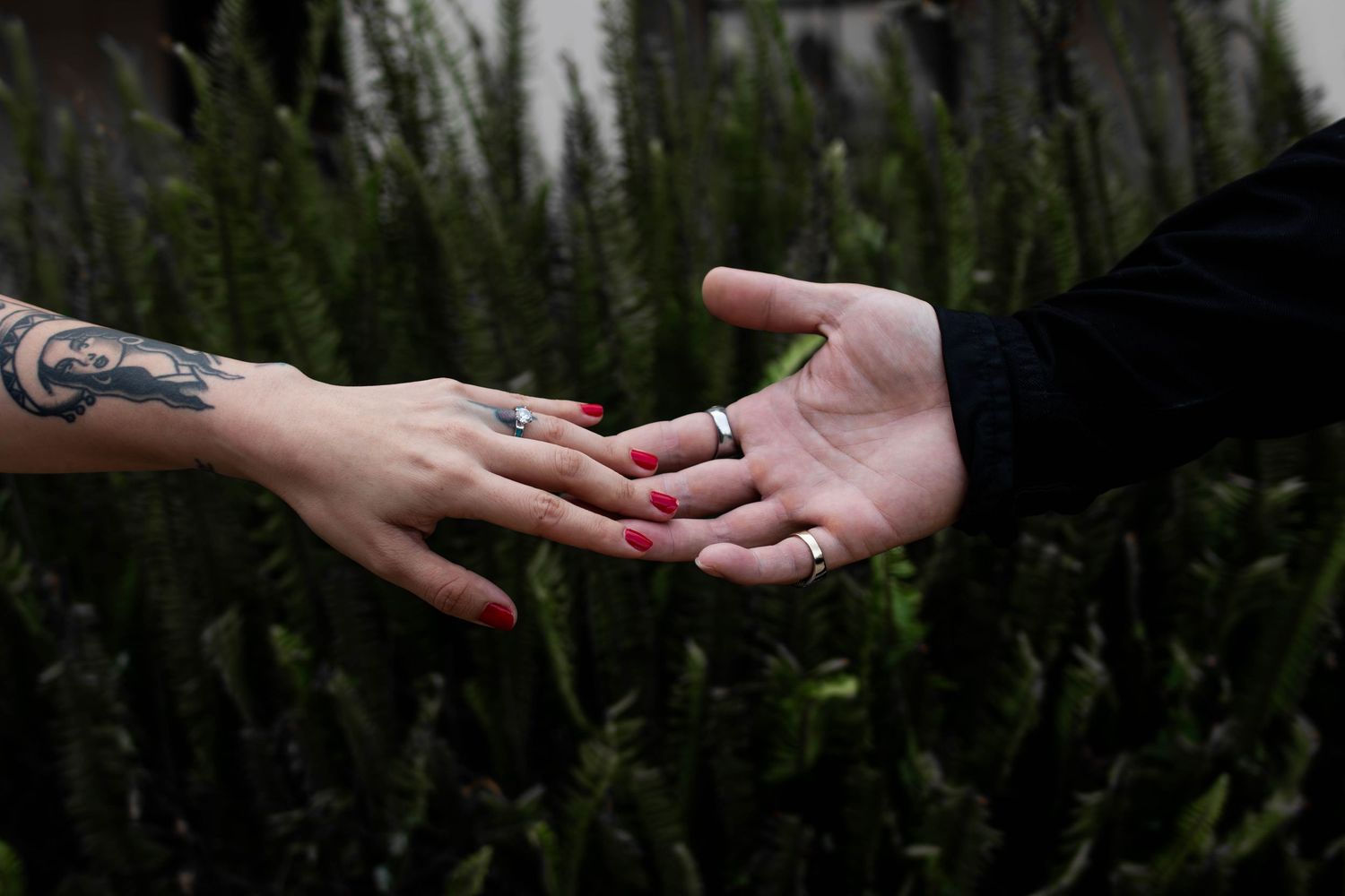 Two hands reaching toward each other, one tattooed with red nails wearing an engagement ring.
