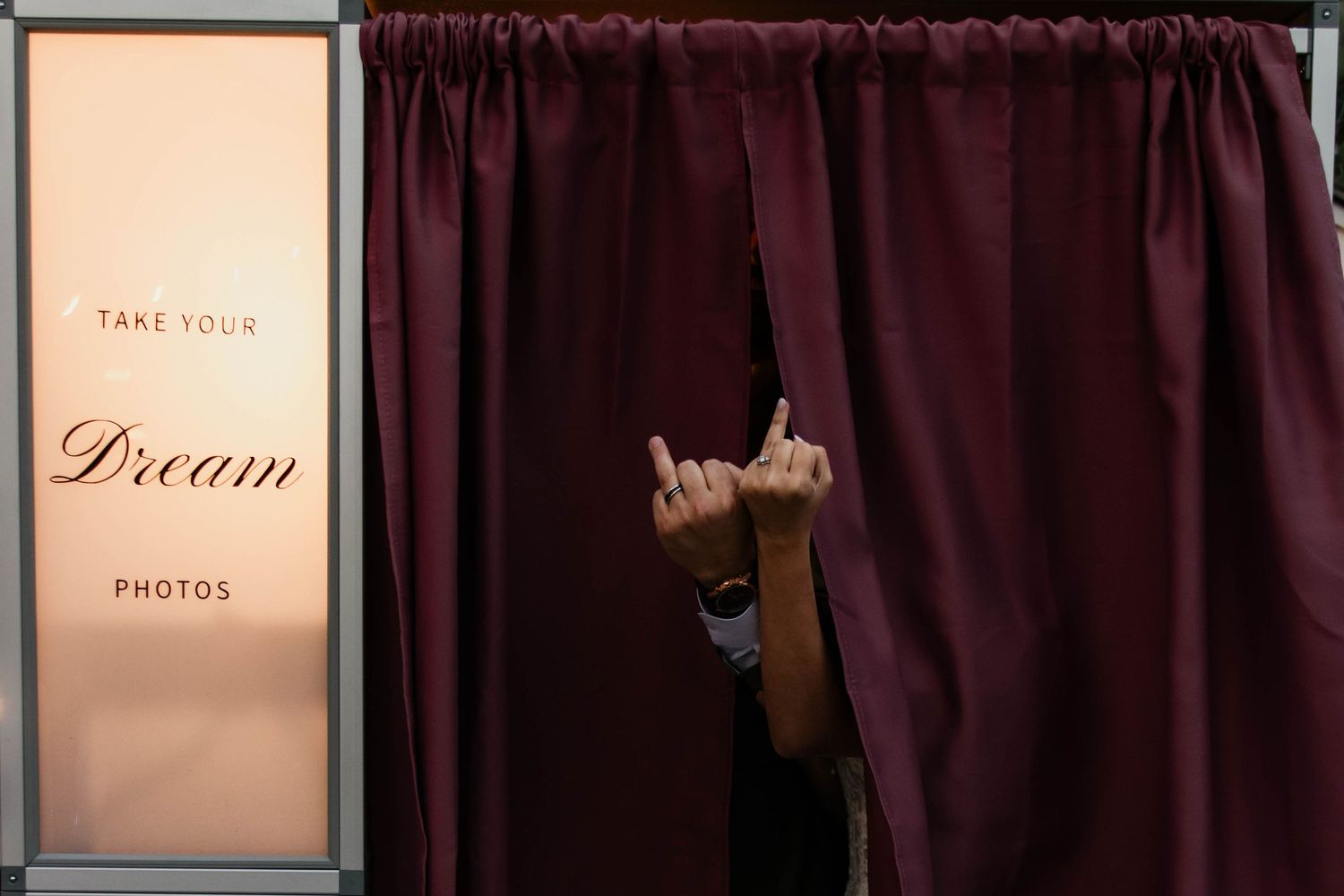 Person peeking through burgundy photo booth curtain making rock sign gesture with hand.