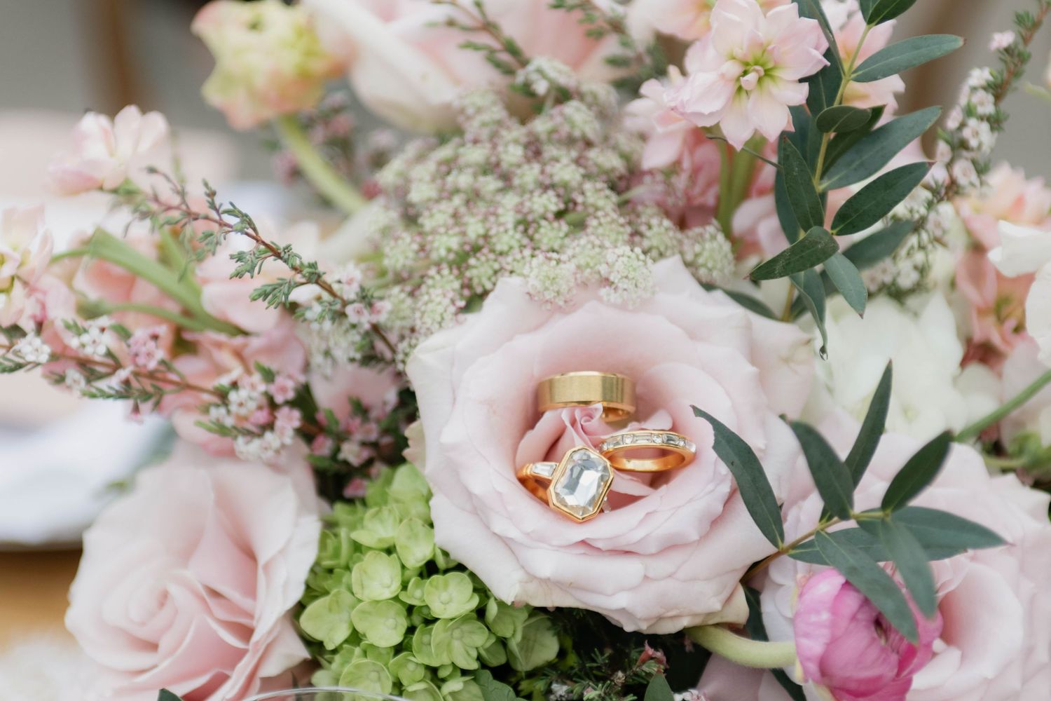 Wedding rings rest elegantly on blush pink roses and greenery in a romantic bridal bouquet.