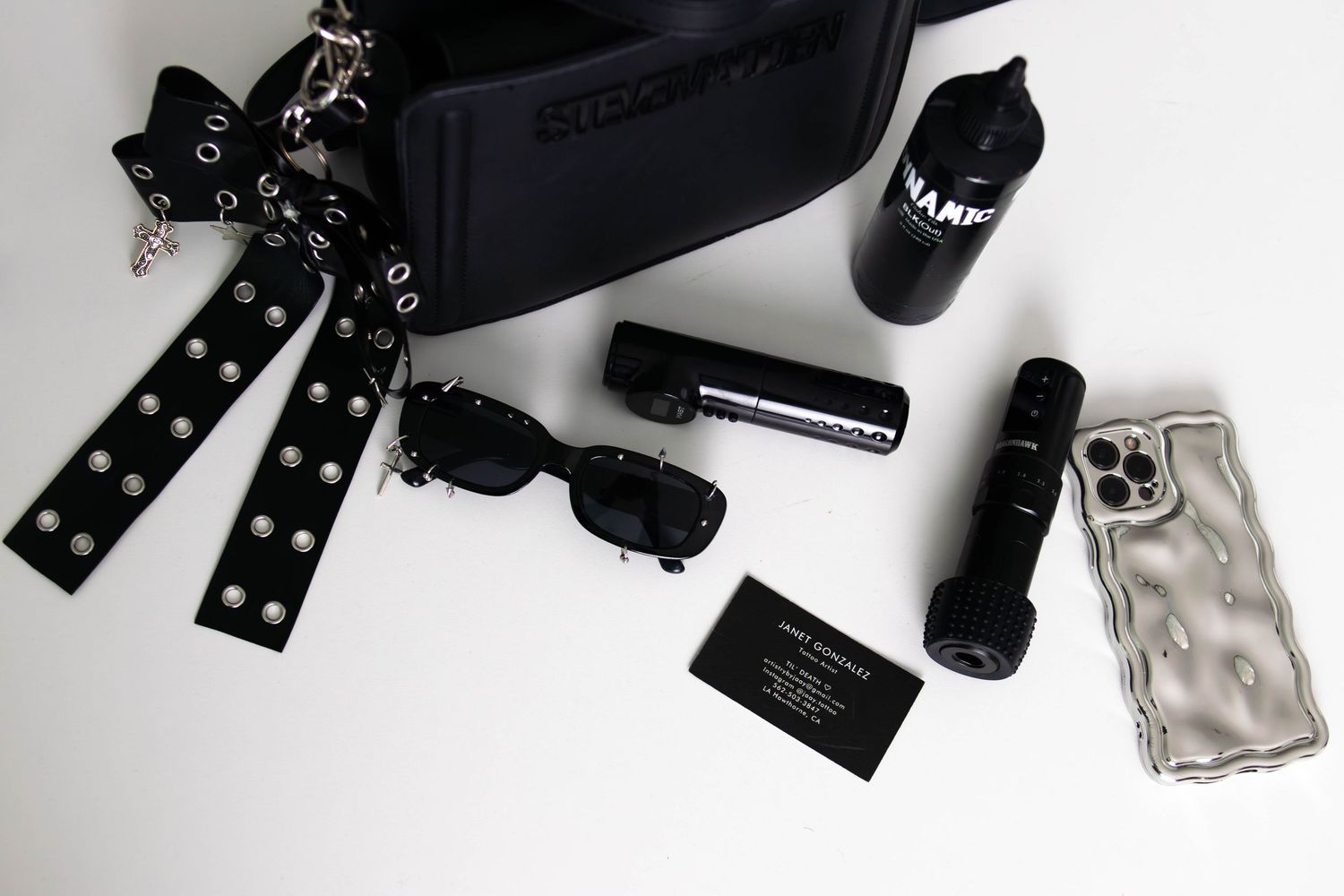 Flatlay of black accessories including sunglasses, bag, water bottle, phone case, and business cards on white background.