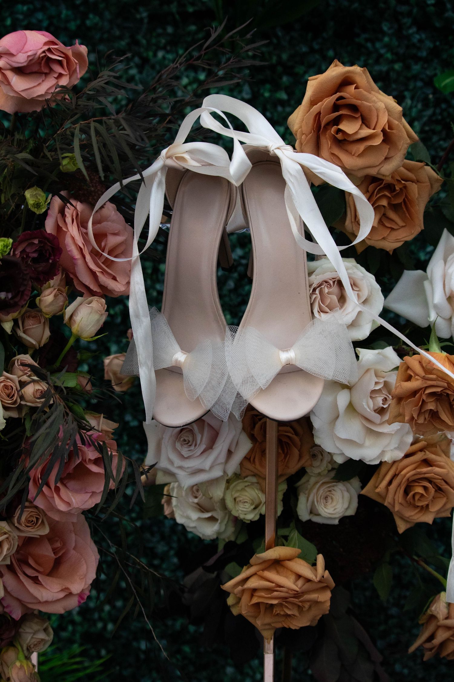 White bridal heels with ribbon laces displayed against a lush arrangement of roses in pink, peach, and burgundy hues.