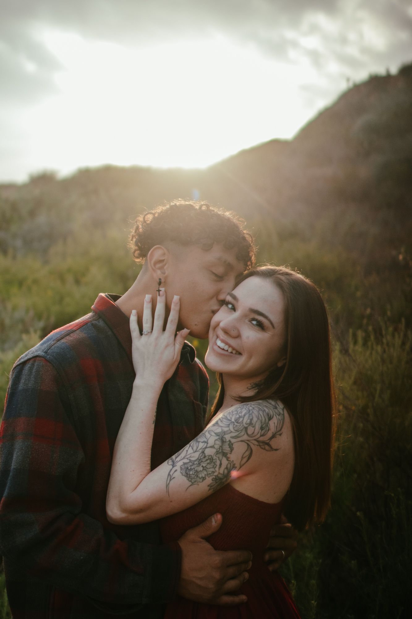 A couple shares a joyful moment with a kiss in a sunlit outdoor mountain setting.