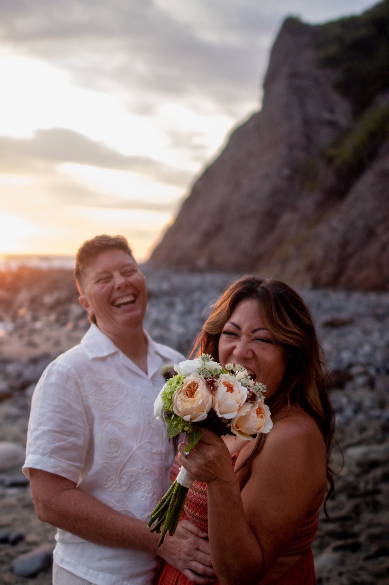 Laughing bride holding white flower bouquet with groom at sunset on rocky beach.