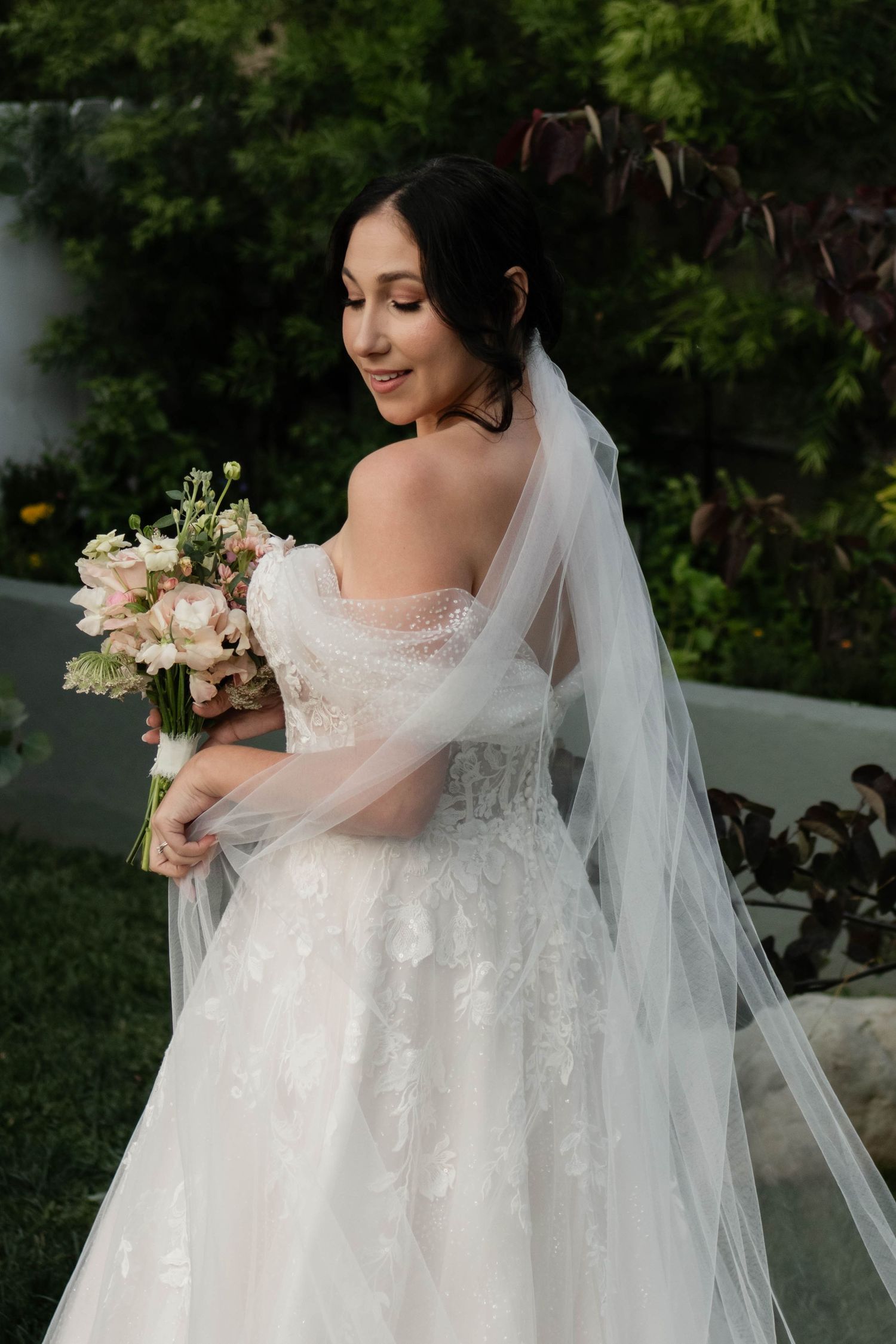 Smiling bride in off-shoulder lace gown with veil holds pink bouquet outdoors in garden setting.