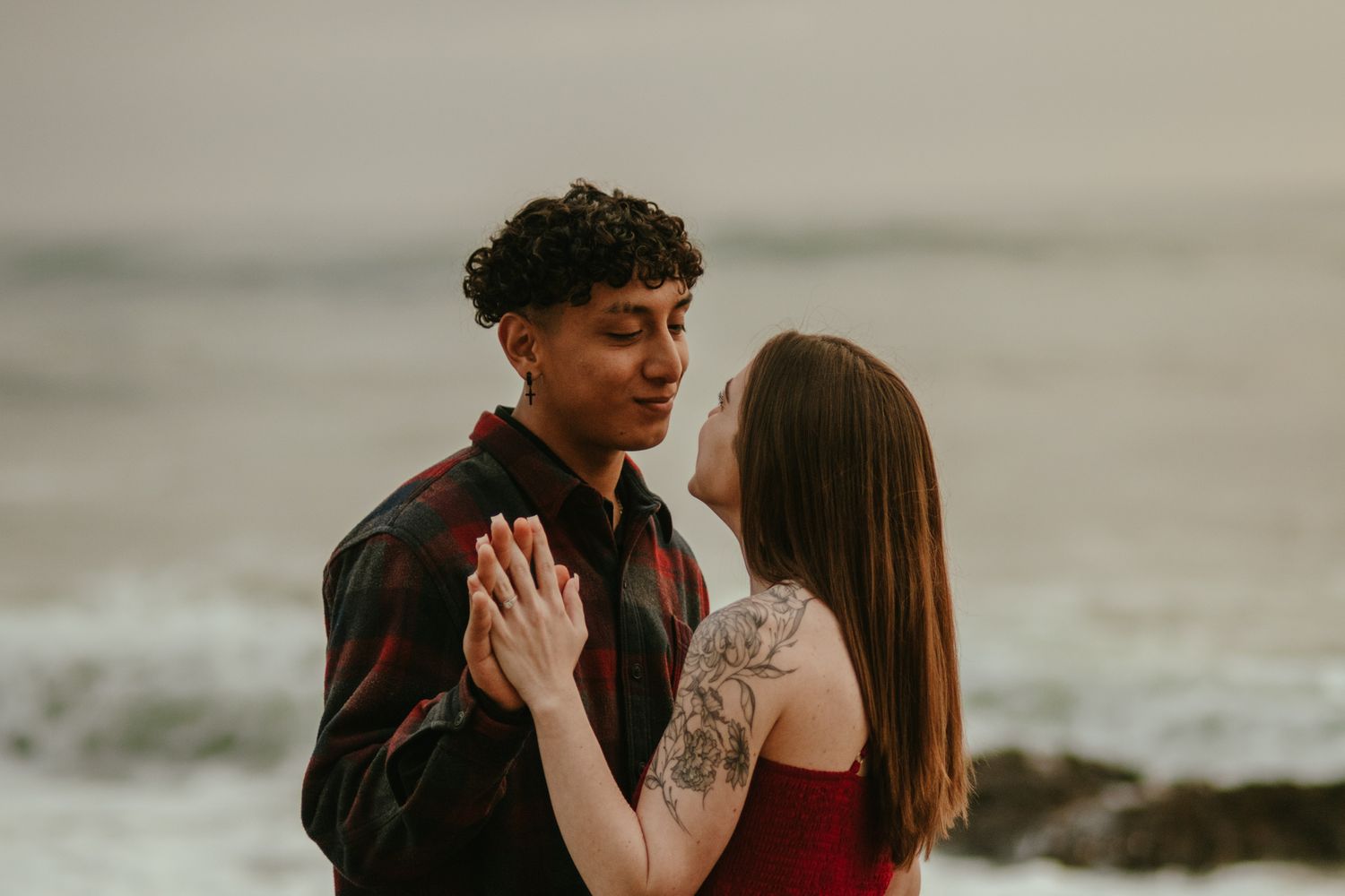 A couple holds hands and gazes at each other near a moody ocean backdrop.