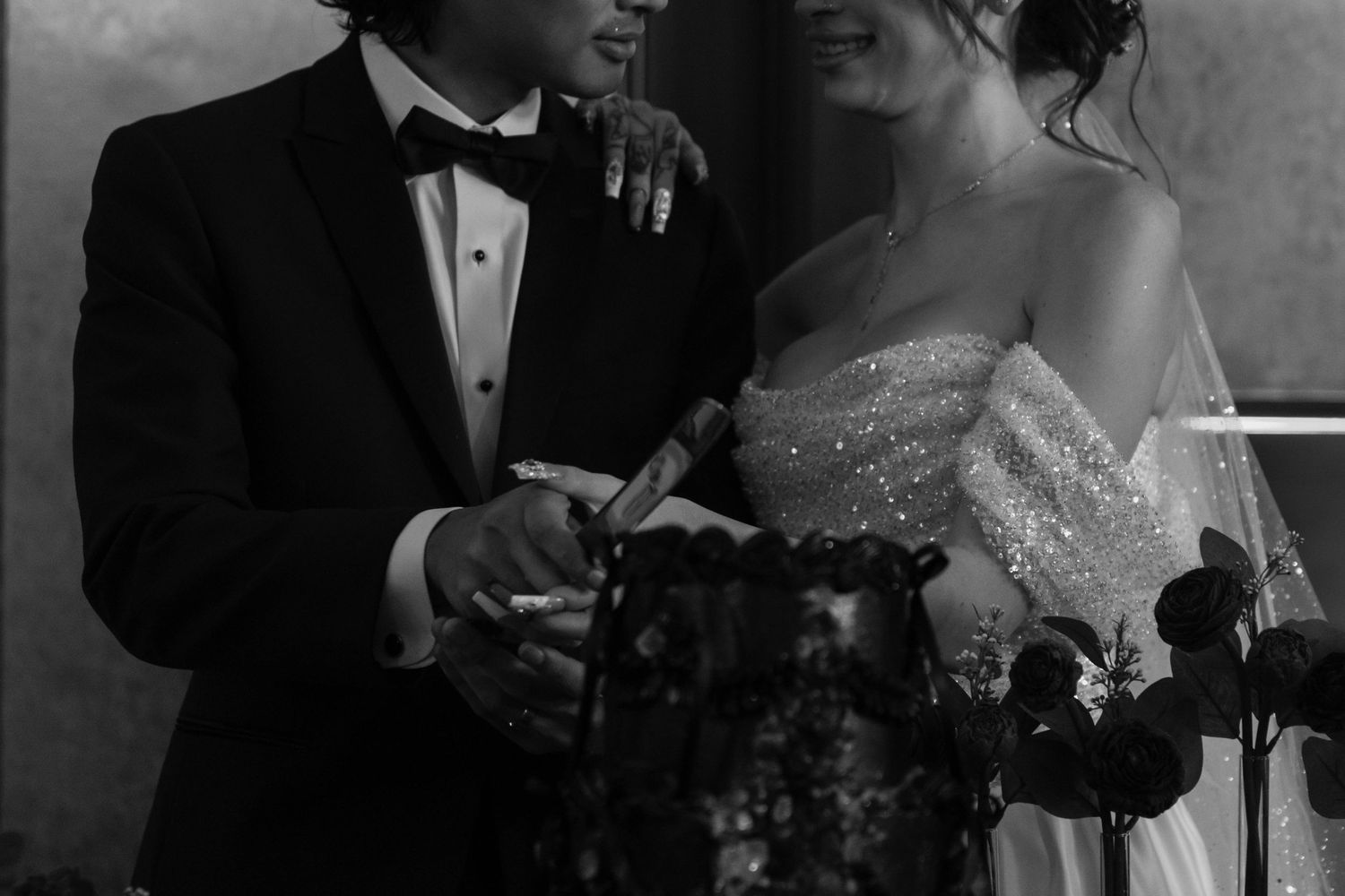 Black and white photo of a bride and groom cutting their wedding cake together in elegant formal attire.