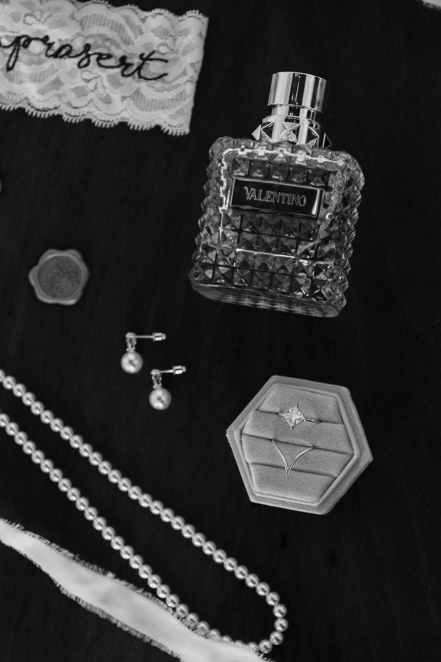 Black and white flat lay of Valentino perfume, pearl necklace, earrings, engagement ring box, and lace wedding accessories.