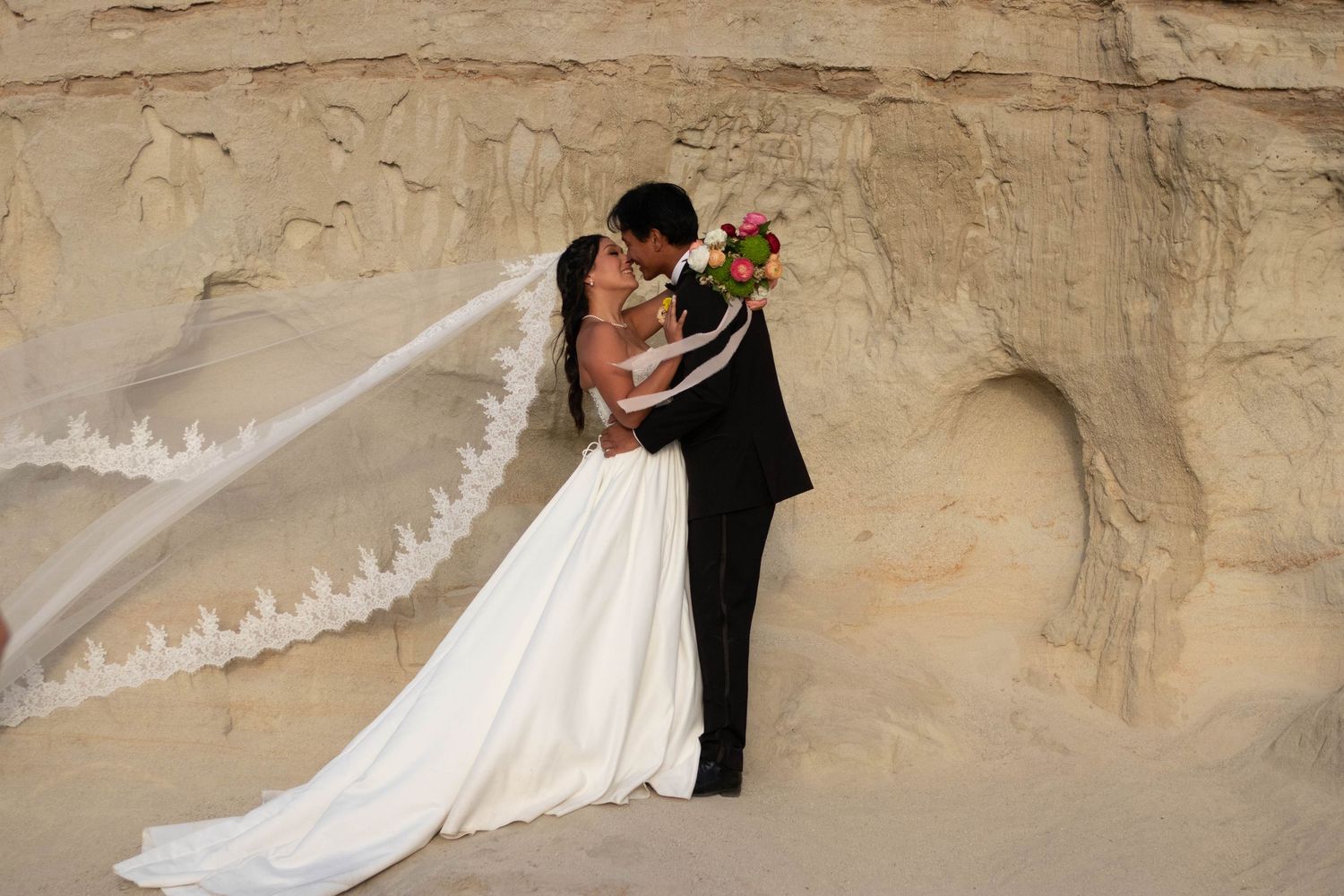 Bride and groom kissing near sandy cliffs, with lace veil flowing in wind, holding colorful bouquet in wedding attire.