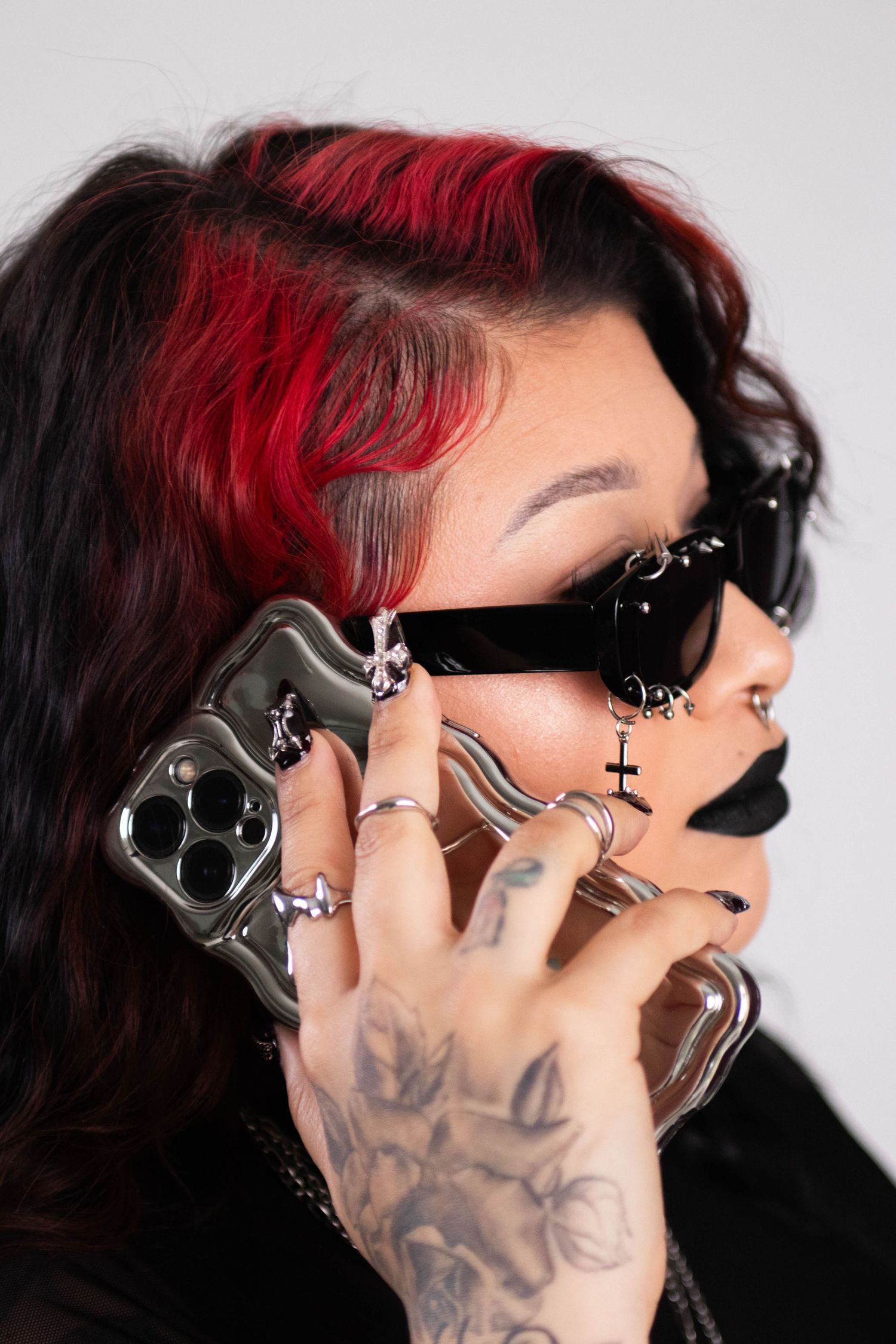 Edgy woman with red and black hair, dark sunglasses, black lipstick, and tattooed hands on phone.