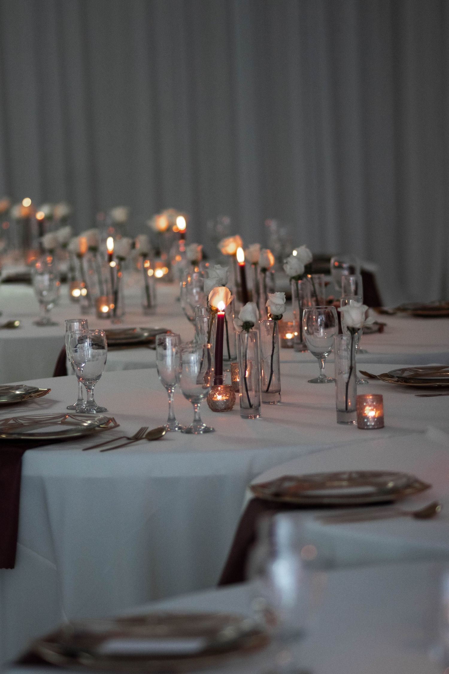 Elegant wedding reception tables set with candles, crystal glasses, and white linens in dim lighting.