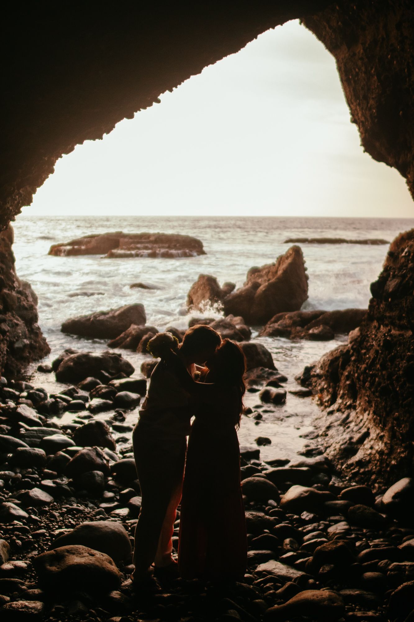 Silhouette of couple kissing inside a sea cave with rocky beach and ocean waves in the background.